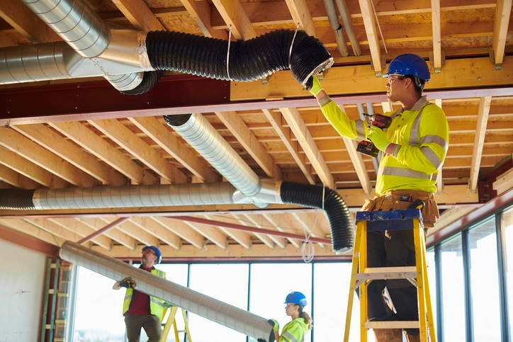 Three construction workers in safety gear and blue helmets install ductwork in a building with exposed wooden beams.