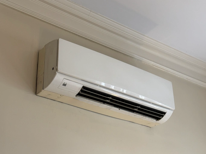 Wall-mounted white air conditioning unit against a light beige wall with white ceiling trim.