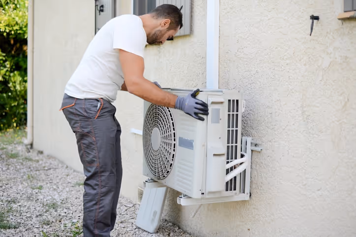 A technician in a white t-shirt and gray pants works on an outdoor air conditioning unit attached to a beige wall