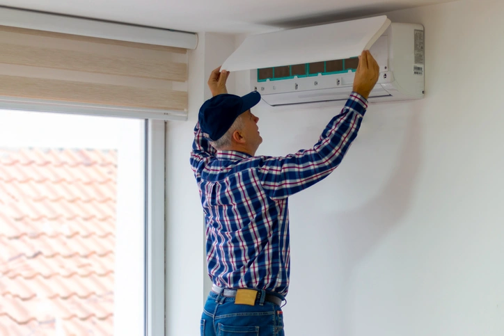 Man in a checked shirt and cap opens air conditioner cover in a bright room.