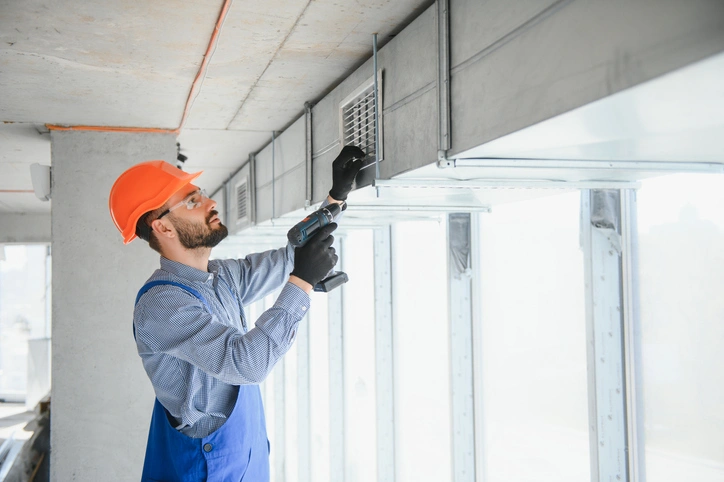 A construction worker in a hard hat and overalls installs a vent using a drill. 