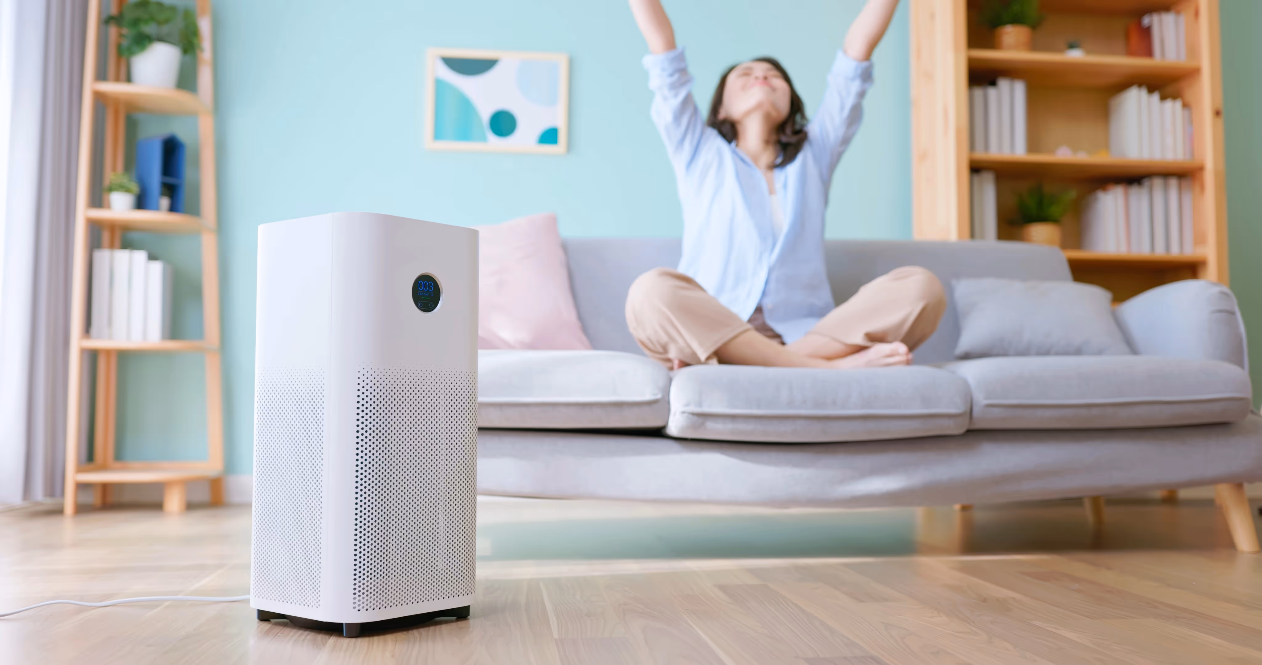 A woman relaxes on a light gray sofa with arms raised in a pastel living room. A white air purifier stands in the foreground, conveying freshness.