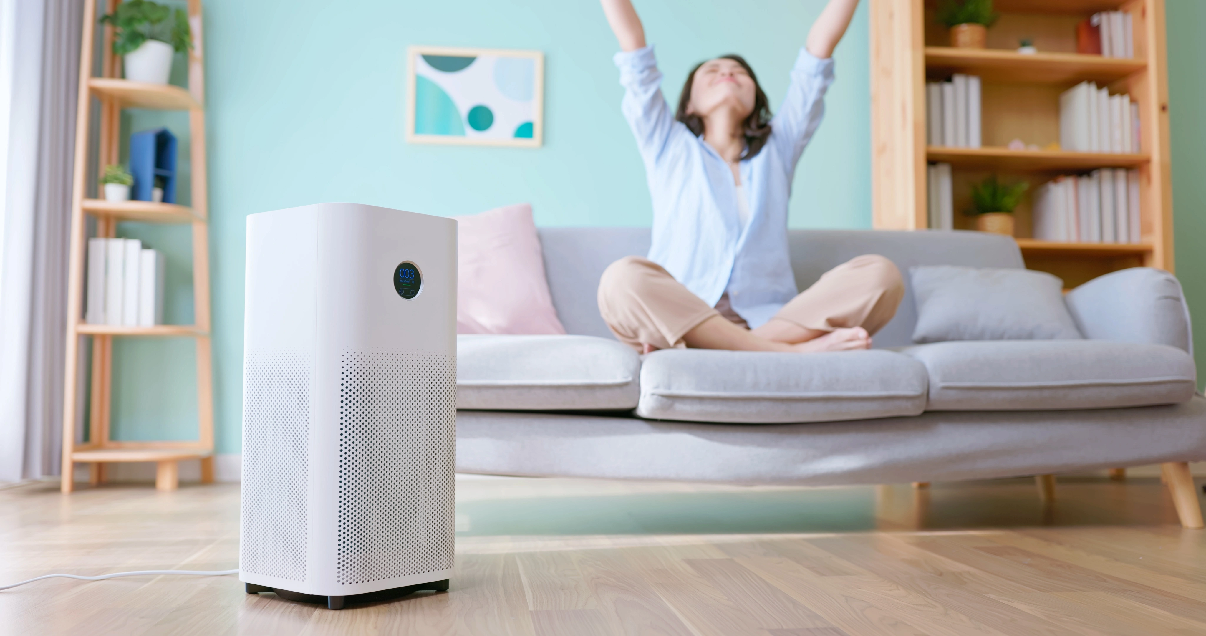A woman relaxes on a light gray sofa with arms raised in a pastel living room. A white air purifier stands in the foreground, conveying freshness.