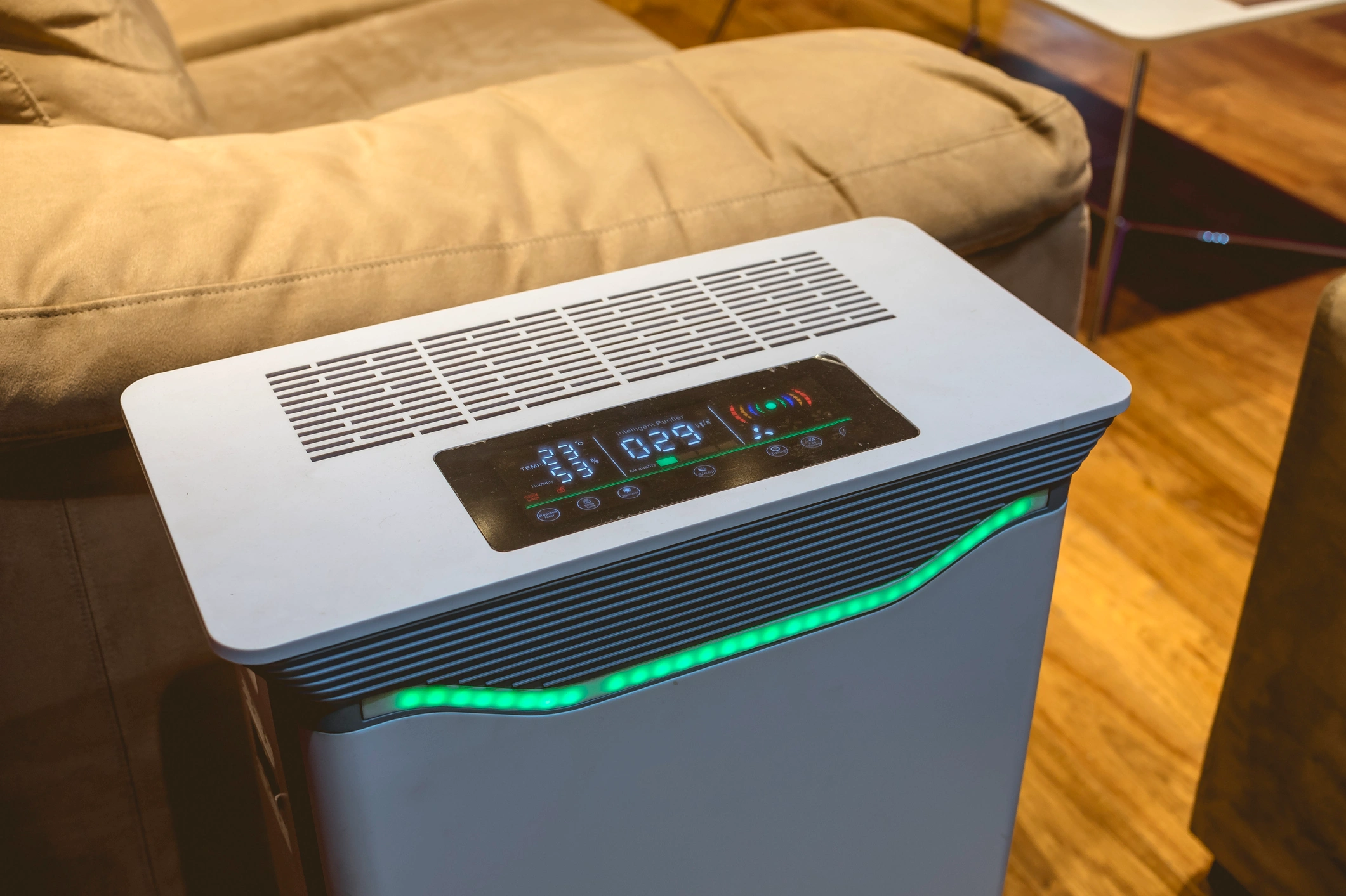 A modern air purifier with a digital display and glowing green lights is placed in a cozy living room with wooden floors and a beige sofa nearby.