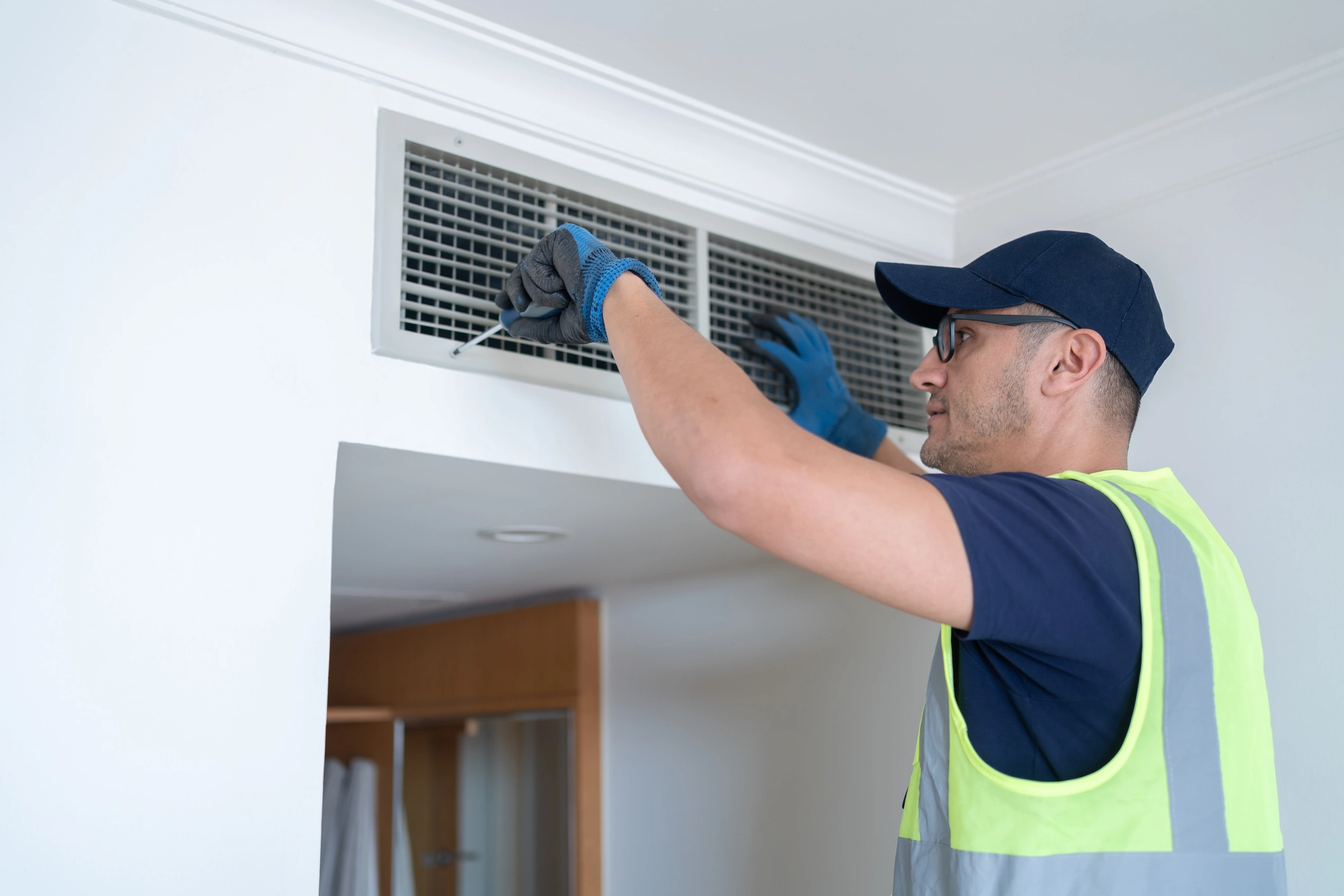 A technician in a neon vest and gloves inspects an air vent on a white wall, focusing intently. 