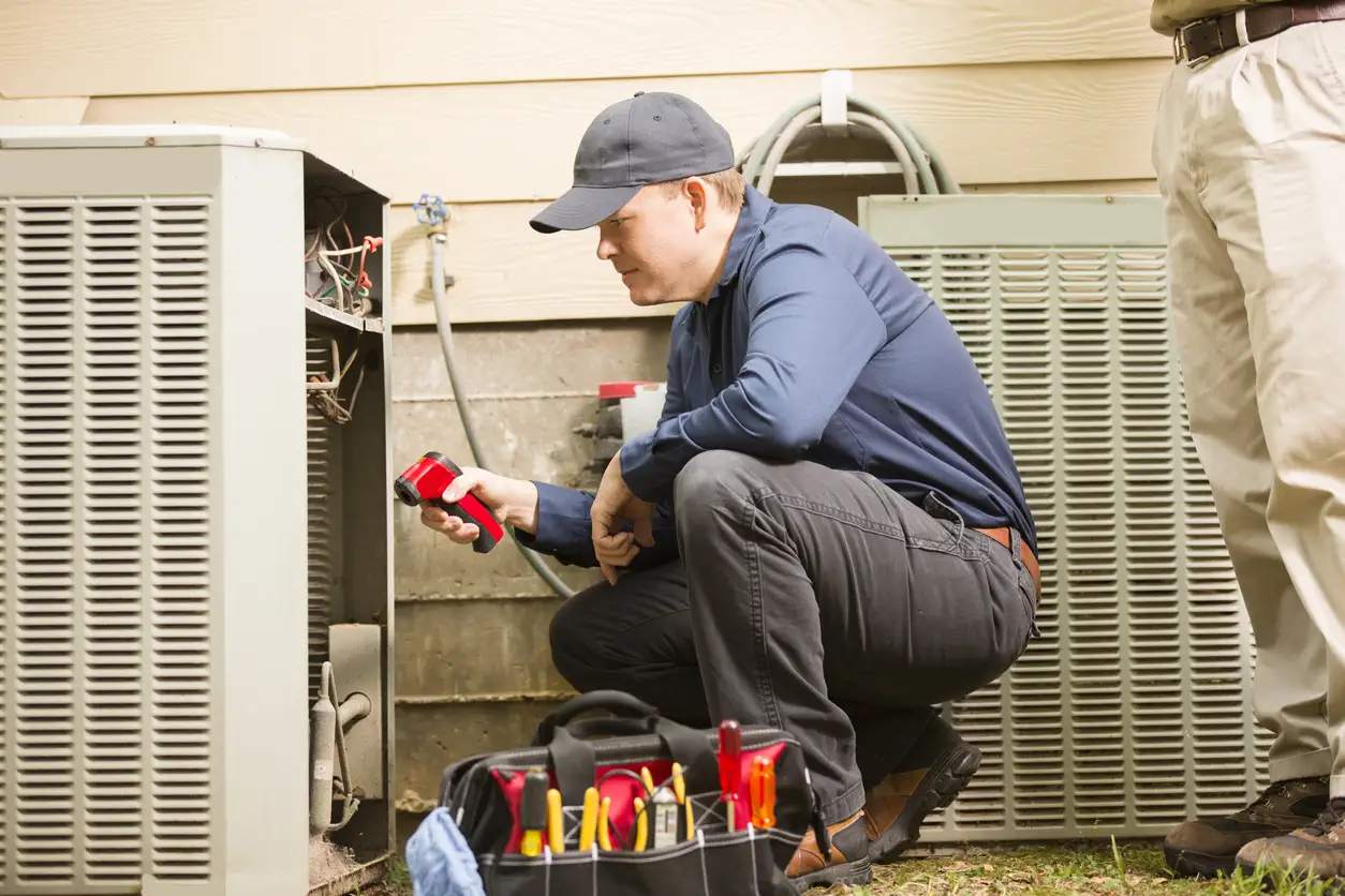 A HVAC service technician is crouched down inspecting the outdoor unit of a heat pump or air conditioner. The technician is holding a thermal imaging camera or thermometer (red and black device) to help diagnose issues, likely checking for temperature anomalies. A tool bag with various tools is open in front of them. Part of another person is visible on the right.