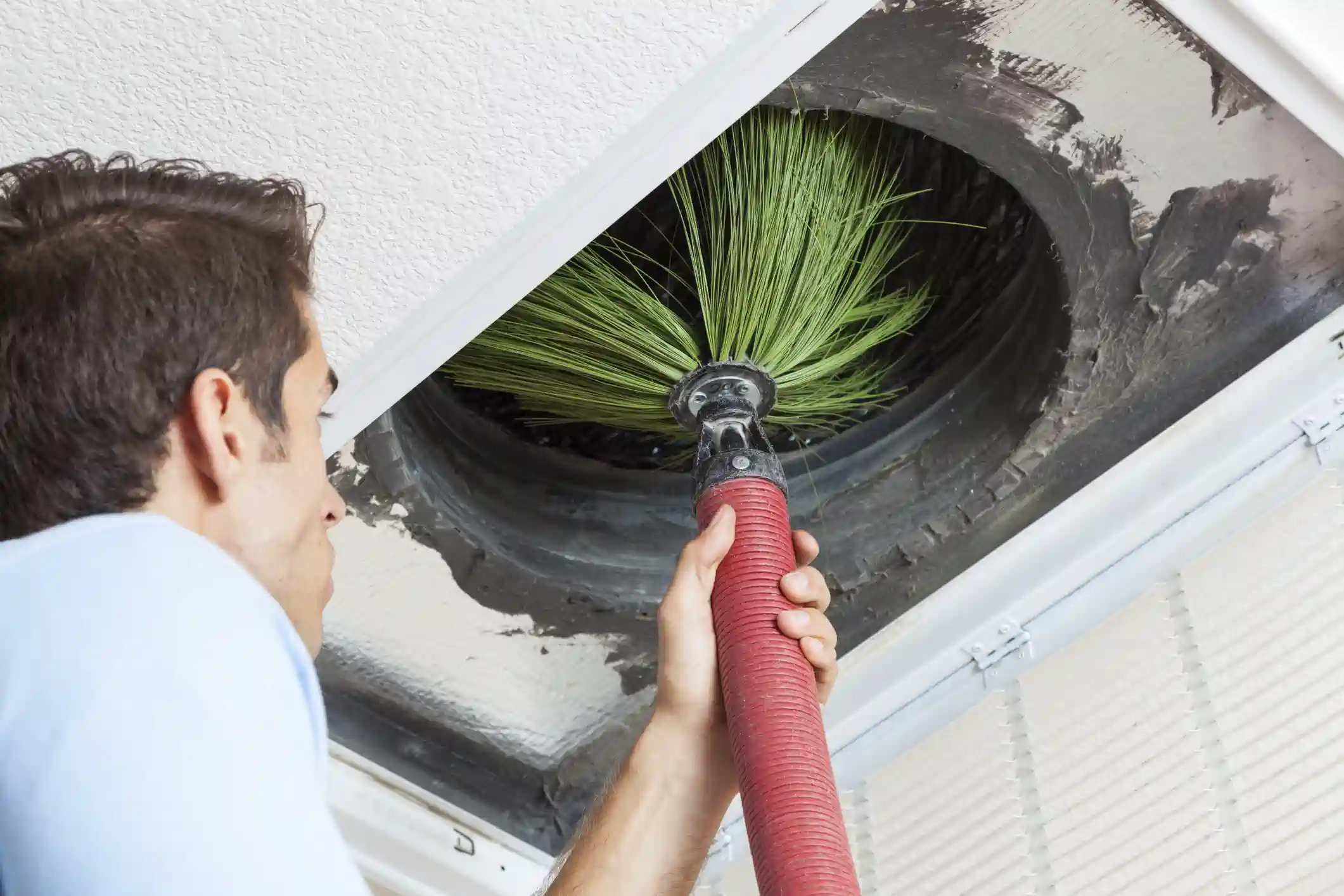  A male technician, seen from the side, is using a large, green, rotating brush attached to a red, flexible hose to clean the interior of an air conditioning vent or duct set into a white ceiling. The opening is square, revealing a circular, dirty duct, illustrating the professional HVAC duct cleaning process.