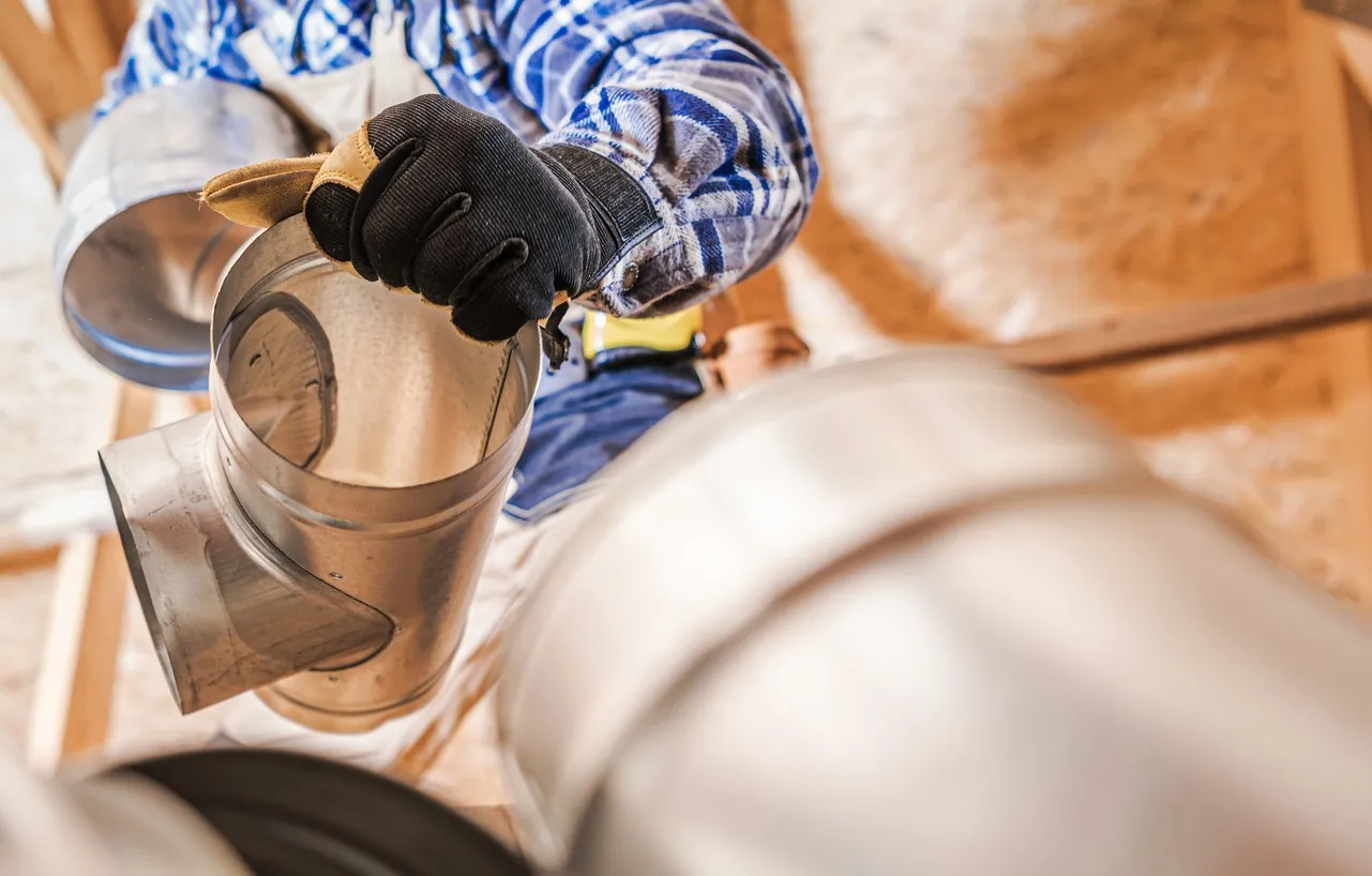  A low-angle close-up shows a technician in a plaid shirt and work gloves reaching up to install a metal Y-connector fitting into a line of cylindrical galvanized ductwork. The perspective is looking straight up at the worker and the ceiling structure, with bare wooden rafters visible in the blurred background of the attic or construction space.