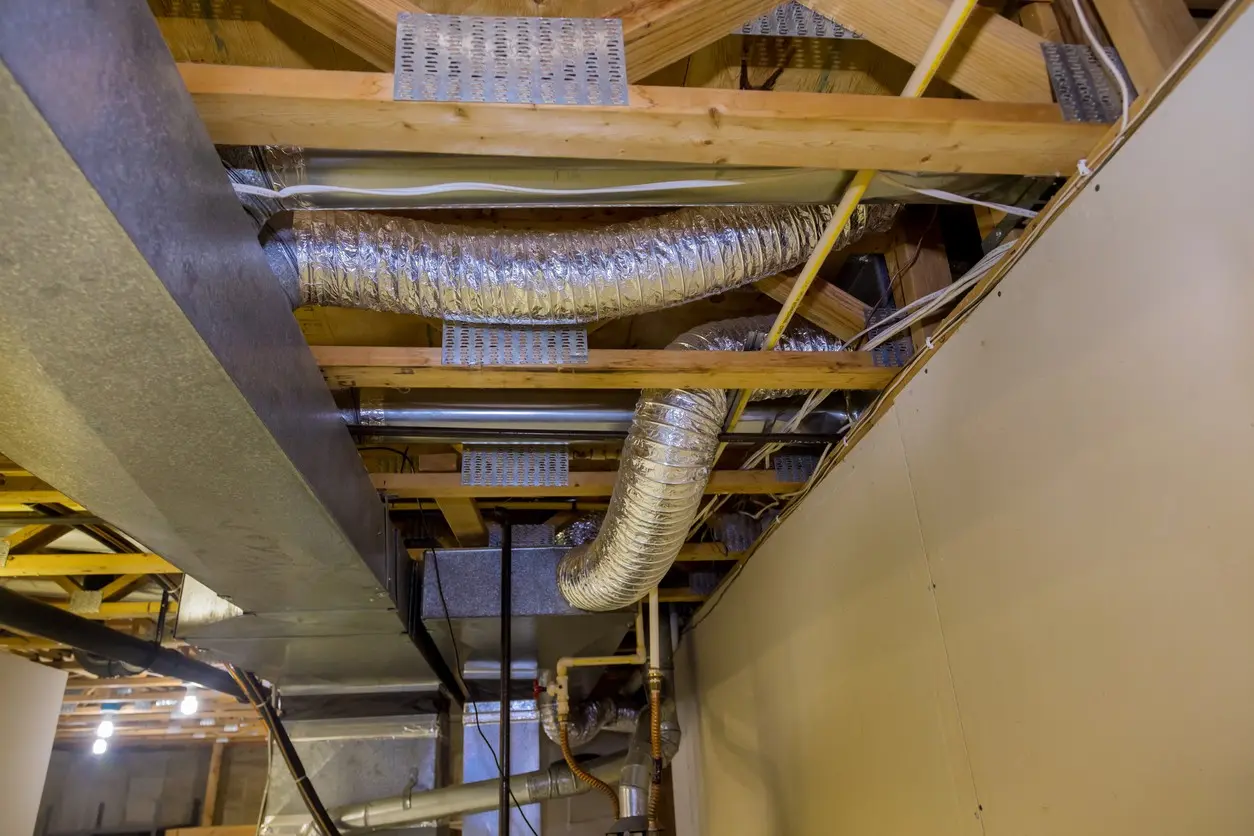  A view from below shows a combination of exposed rigid rectangular galvanized metal ductwork and flexible, foil-wrapped insulated ductwork running through wooden floor joists in a residential basement or unfinished area. Metal truss plates connect the wooden beams, and various wires and pipes are also visible among the HVAC components.