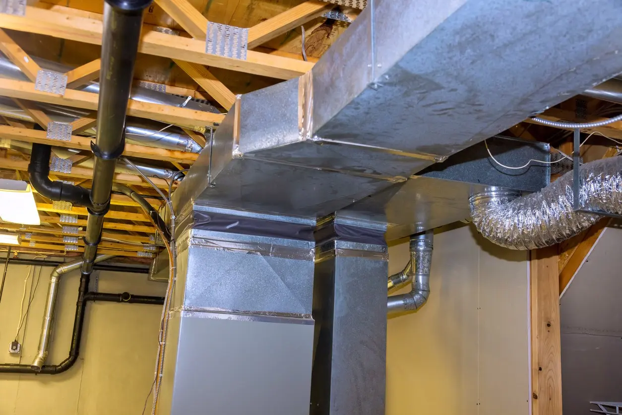 A view of exposed ductwork in a basement or utility room shows a large central rectangular galvanized metal duct descending from the ceiling, likely the main HVAC supply or return plenum. It connects to horizontal rectangular trunk lines and smaller flexible foil-wrapped ducts. The ceiling structure, including wooden joists and metal plates, along with various black and silver plumbing pipes, are visible overhead.