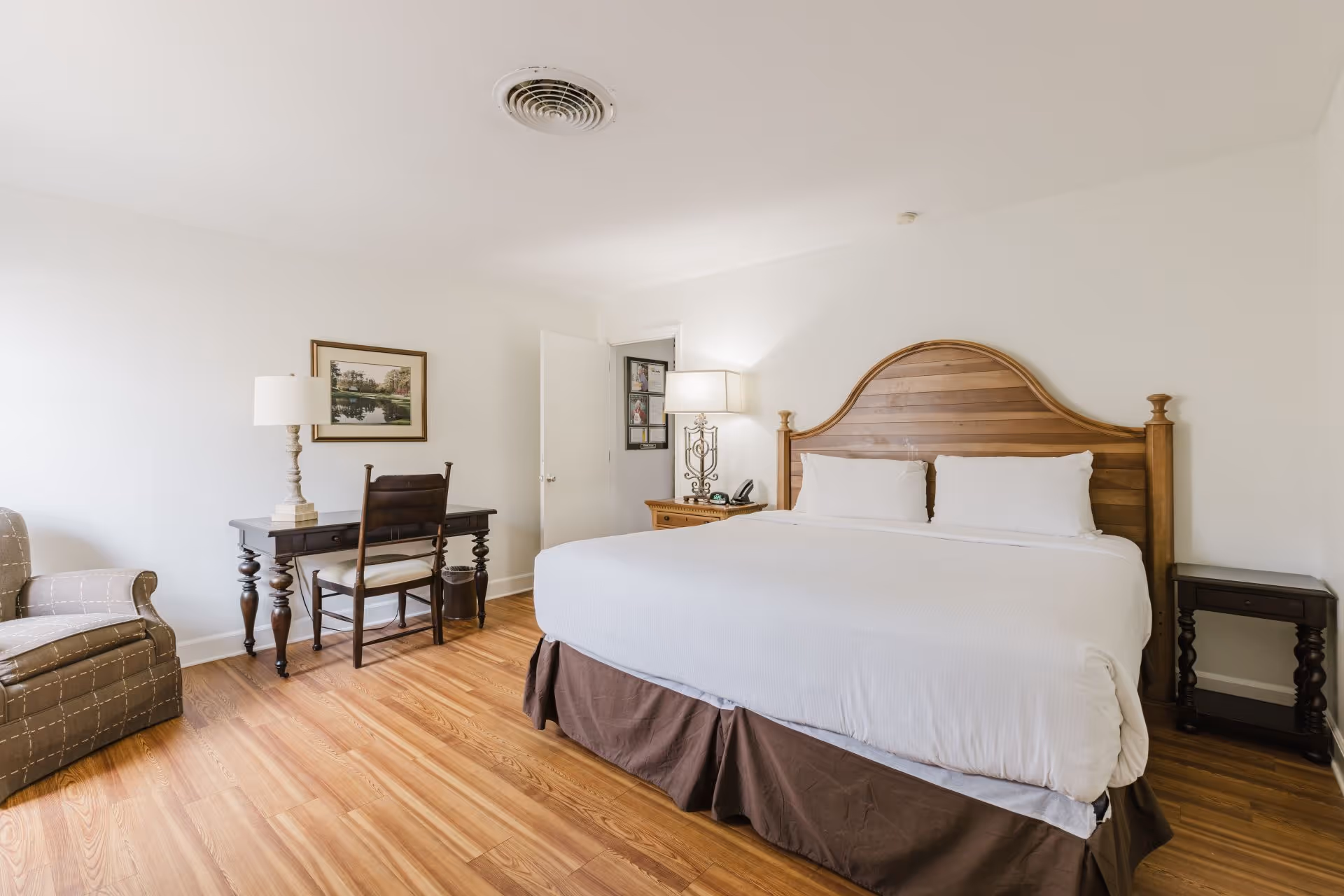 Bright hotel room with a large wooden headboard king bed, a desk with chair and lamp, an armchair, and wooden flooring.