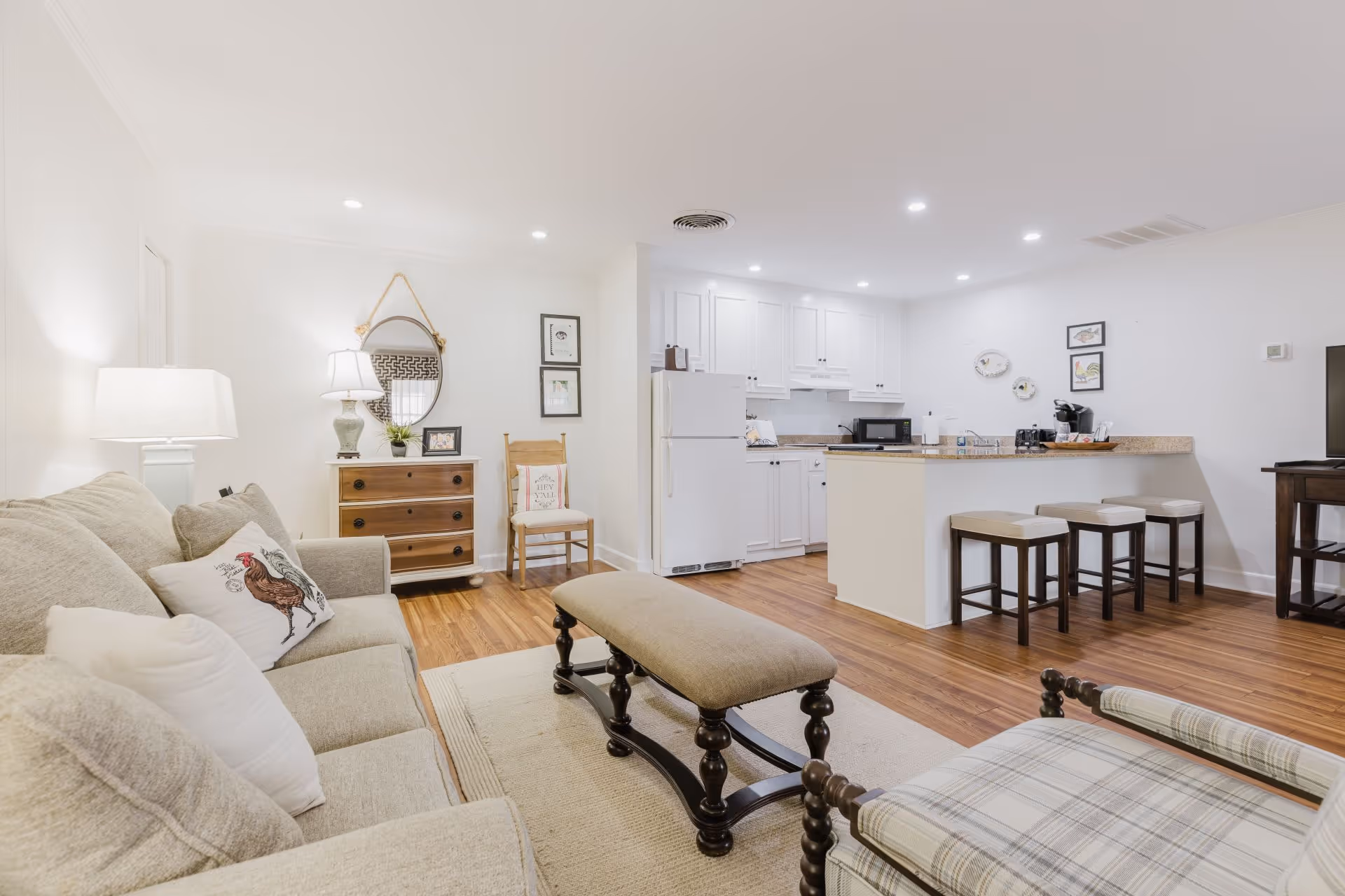 Bright living space with beige sofa, wooden bench, plaid armchair, small kitchen with white cabinets and breakfast bar with three stools.