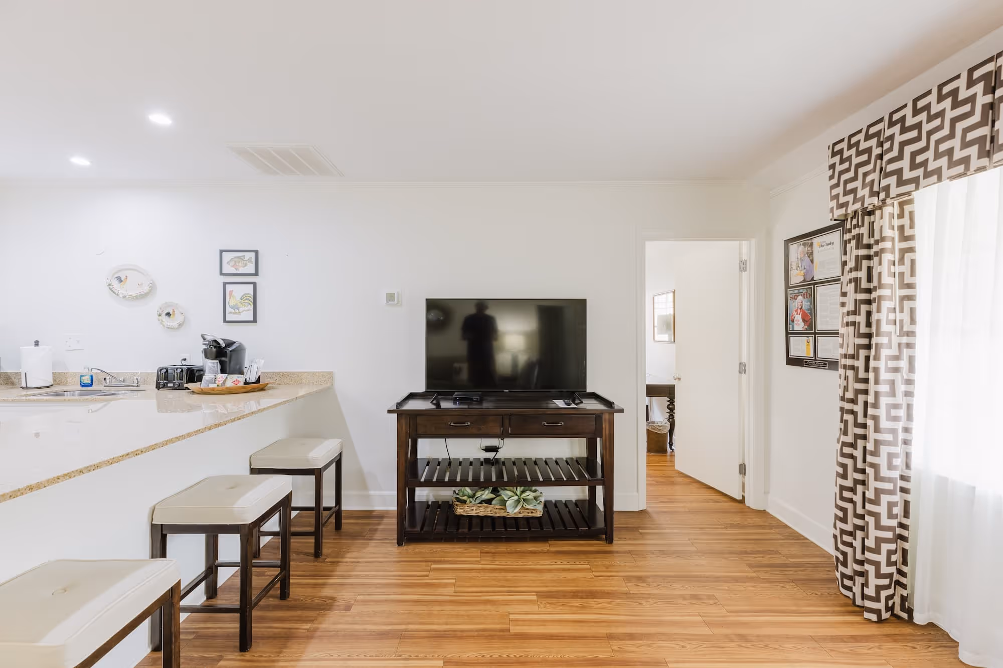 Bright living space with a flat-screen TV on a dark wood console, beige bar stools, and patterned brown and white curtains by a window.
