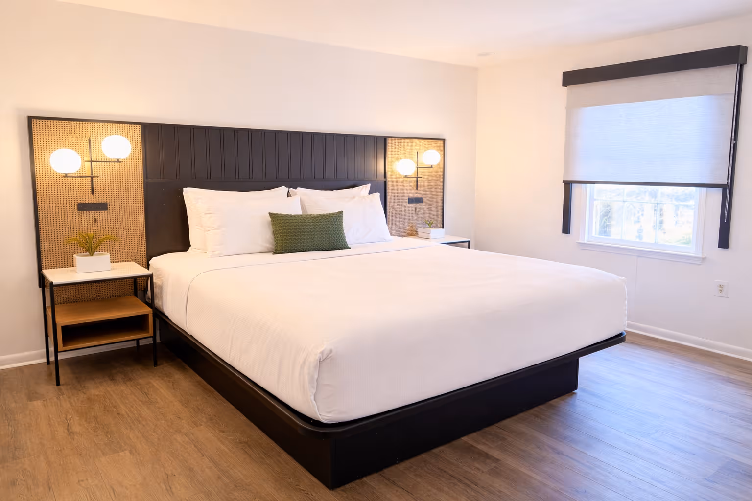 Modern bedroom with large bed featuring white linens, a green decorative pillow, wooden floor, and wall-mounted globe lights on rattan panels.