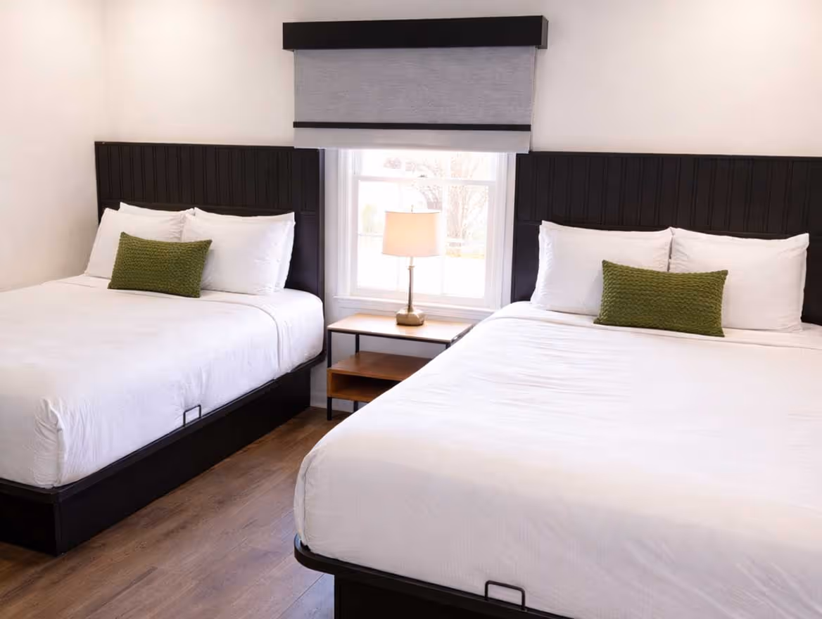 Hotel room with two beds featuring white linens, green accent pillows, wood floors, and a nightstand with a lamp in front of a window with a gray shade.