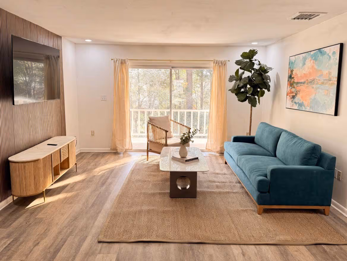 Bright living room with teal sofa, beige armchair, large rug, modern coffee table, flat-screen TV, and a large window with curtains.