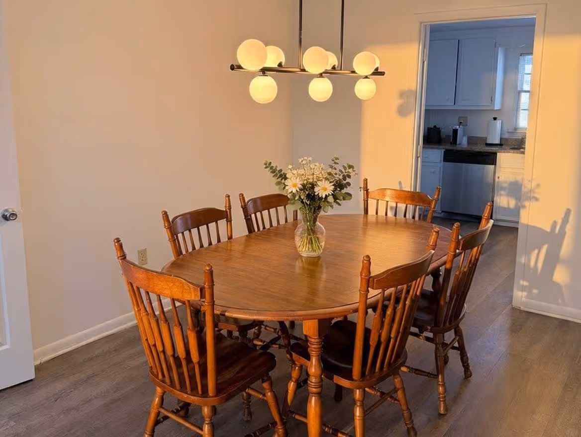 Wooden oval dining table with six matching chairs, a vase of flowers in the center, and a modern chandelier above in a dining room.