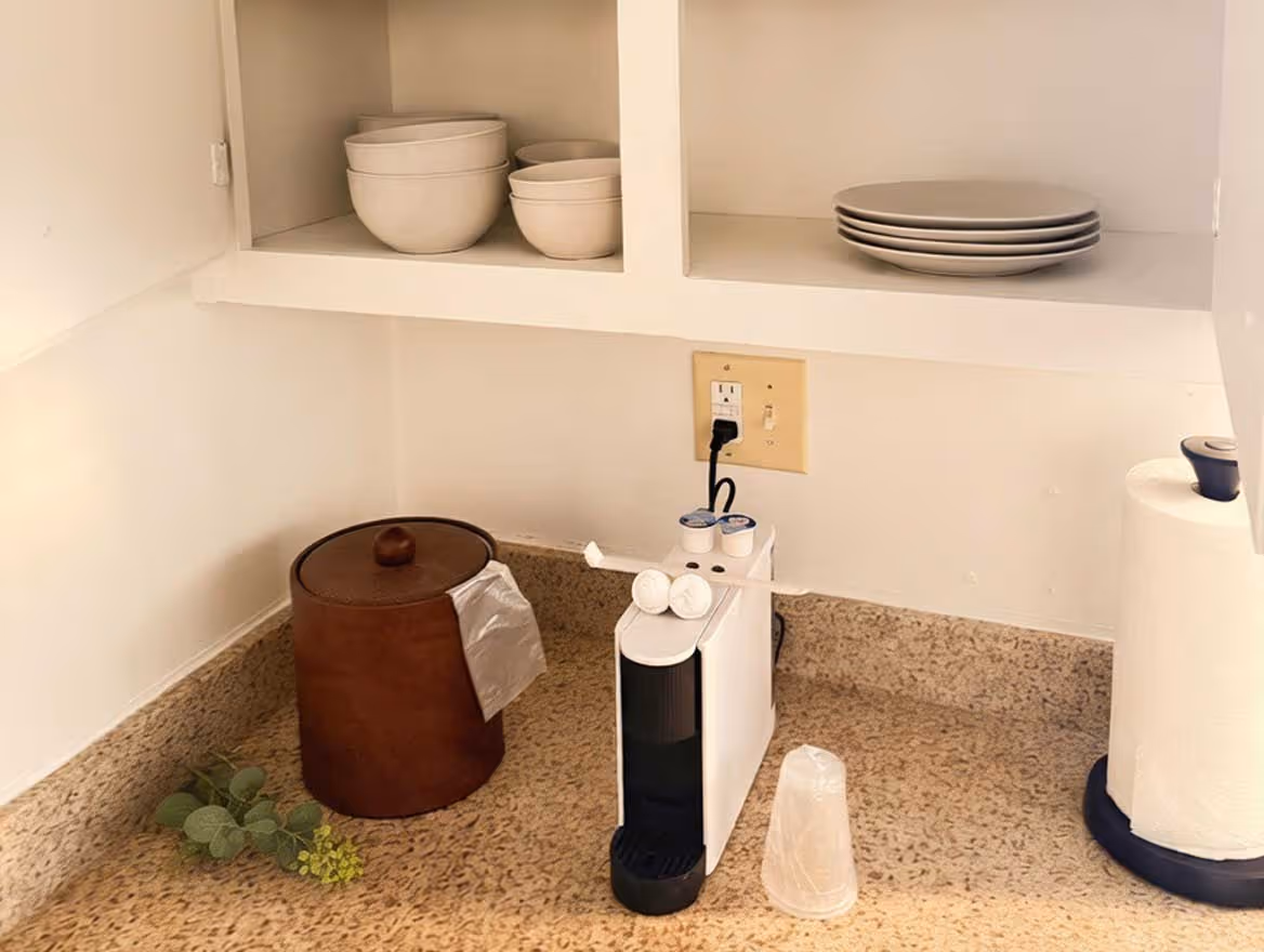 Kitchen countertop with a white coffee pod machine, brown container with a lid, stacked white bowls and plates on open shelves, paper towel roll, and a small plant sprig.