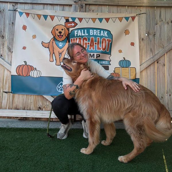 Camp counselor hugging a golden retriever in front of the Fall Break Wag-A-Lot Camp banner