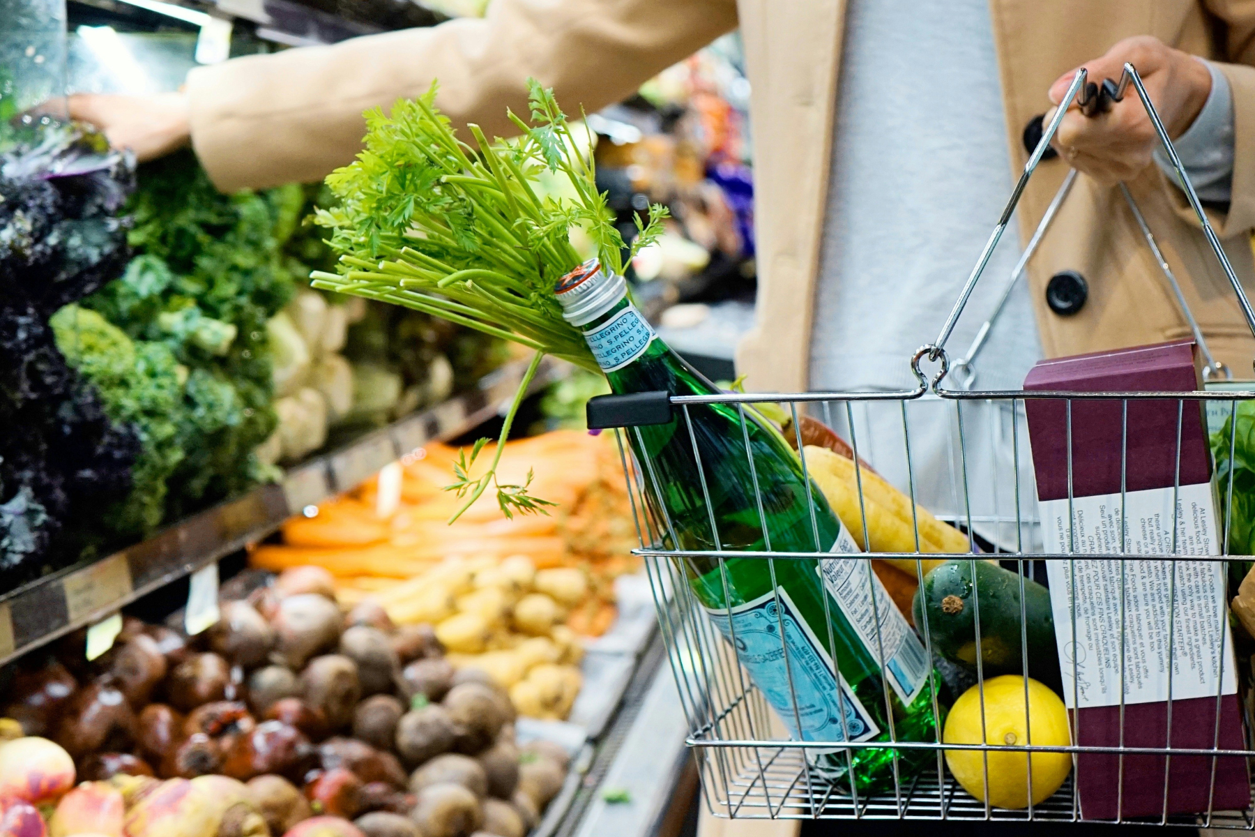 A close-up of a person holding a shopping basket in a supermarket, reaching for vegetables