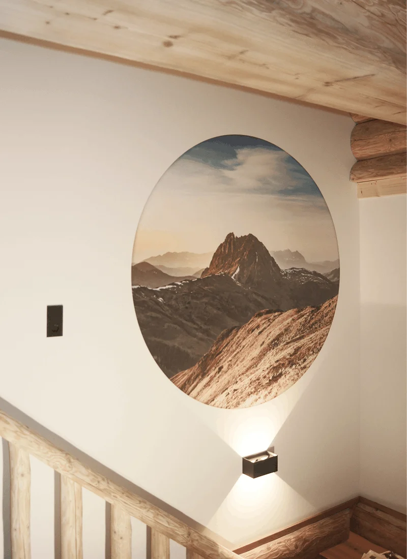 Rundes Wandbild einer Berglandschaft über einer Treppe mit Holzgeländer, beleuchtet von einer Wandlampe.