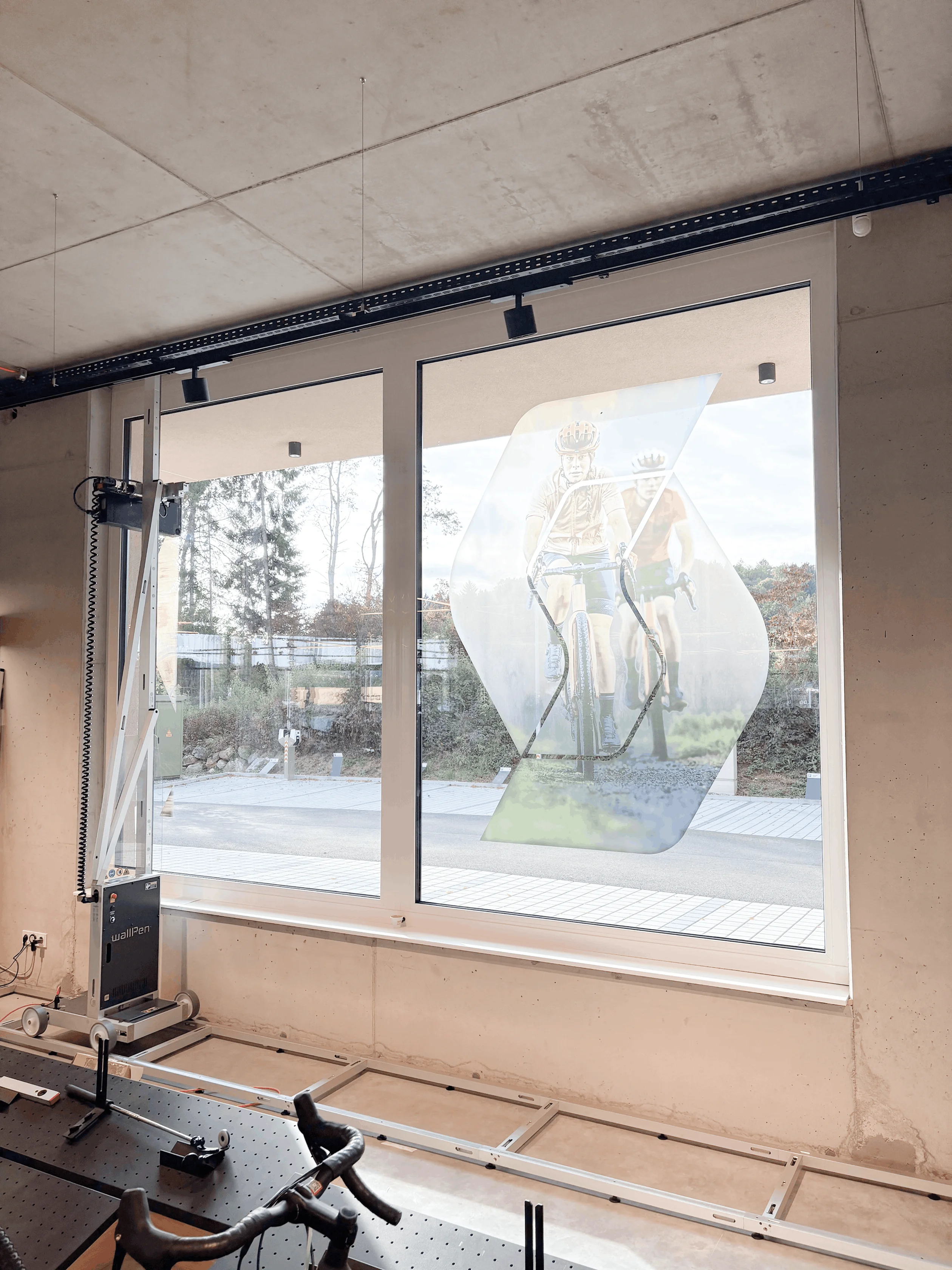 Maschine zeichnet ein Bild von zwei Fahrradfahrern auf ein großes Fenster in einem industriellen Innenraum.
