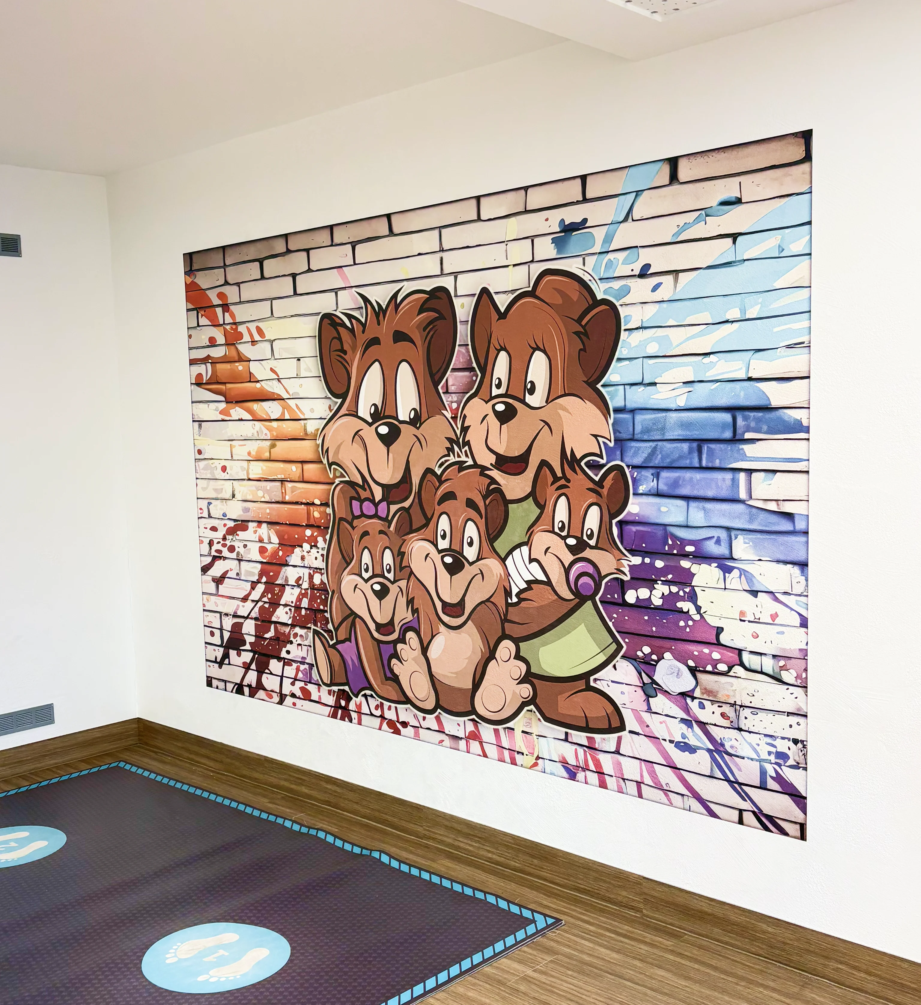 Wandbild mit fünf fröhlichen Comic-Bären vor einer bunt gesprayten Ziegelsteinwand in einem hellen Raum mit Holzboden.