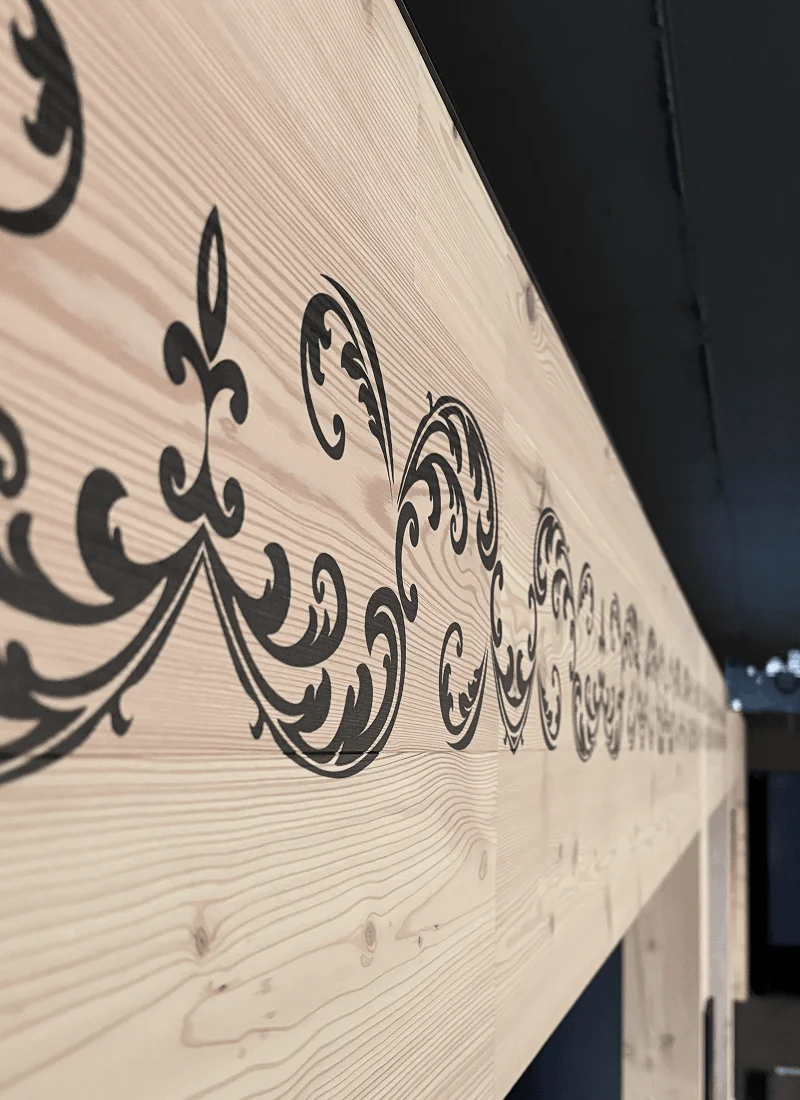 Holzpaneel mit schwarzem, dekorativem, geschwungenem Muster vor dunklem Hintergrund.