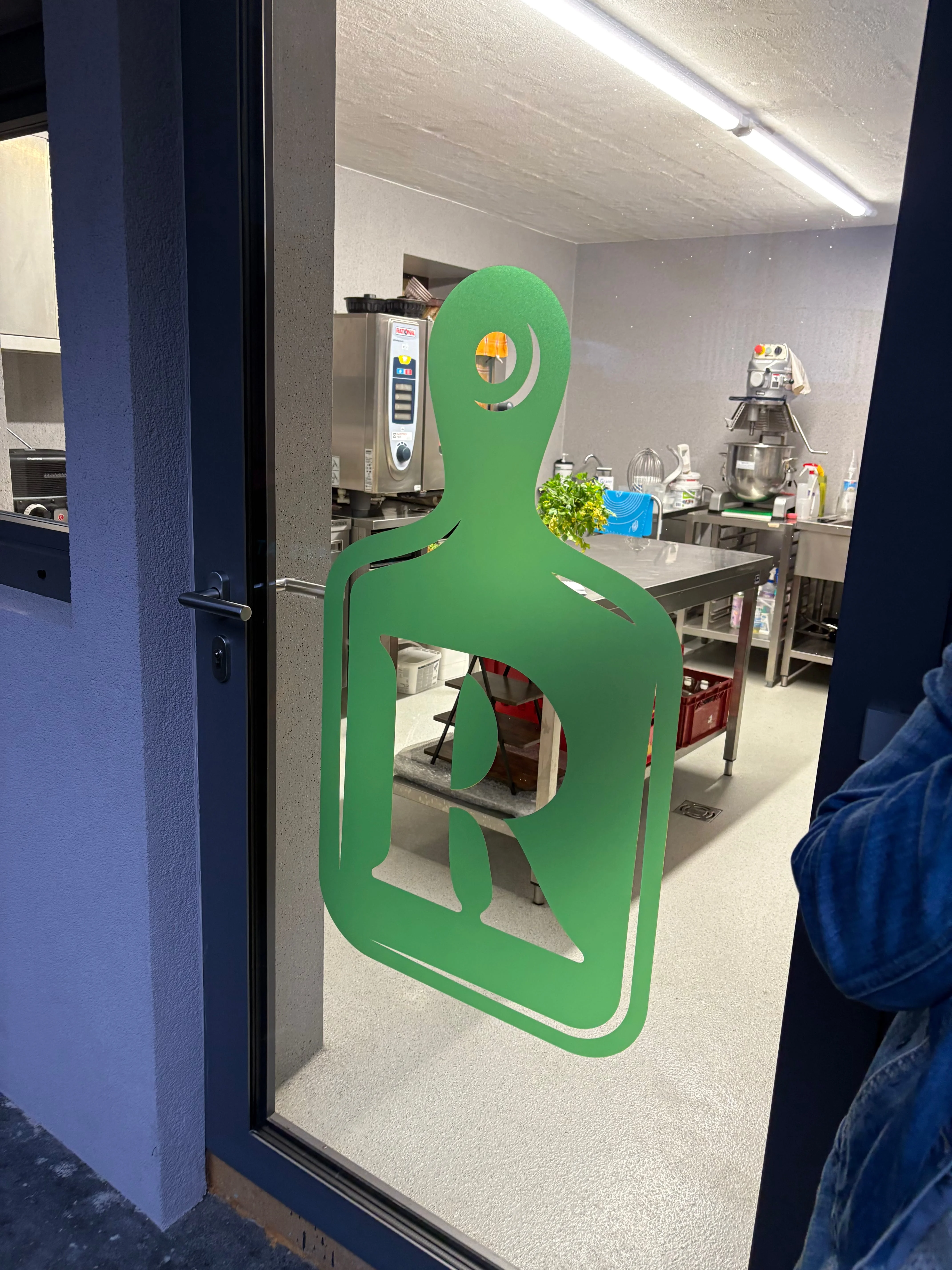 Grünes Logo mit stilisiertem Buchstaben R auf einer Glastür, Blick in eine moderne Küche mit Edelstahl-Arbeitstischen und Küchenmaschinen.