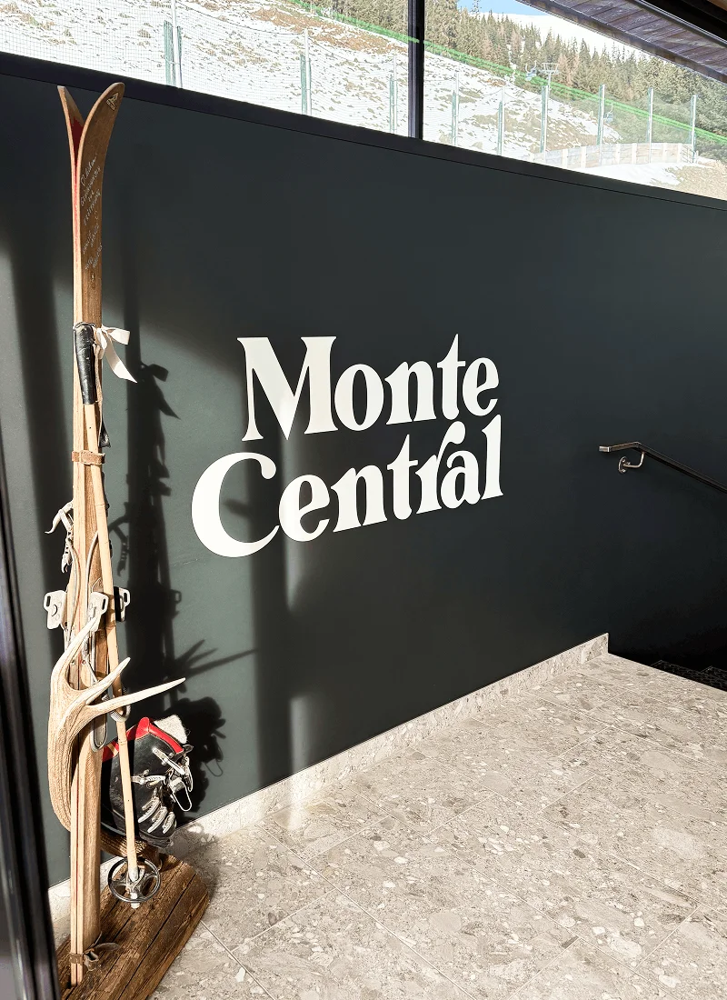 Holzskier, Skistöcke und ein Paar Skischuhe stehen neben einer schwarzen Wand mit dem Schriftzug 'Monte Central'.