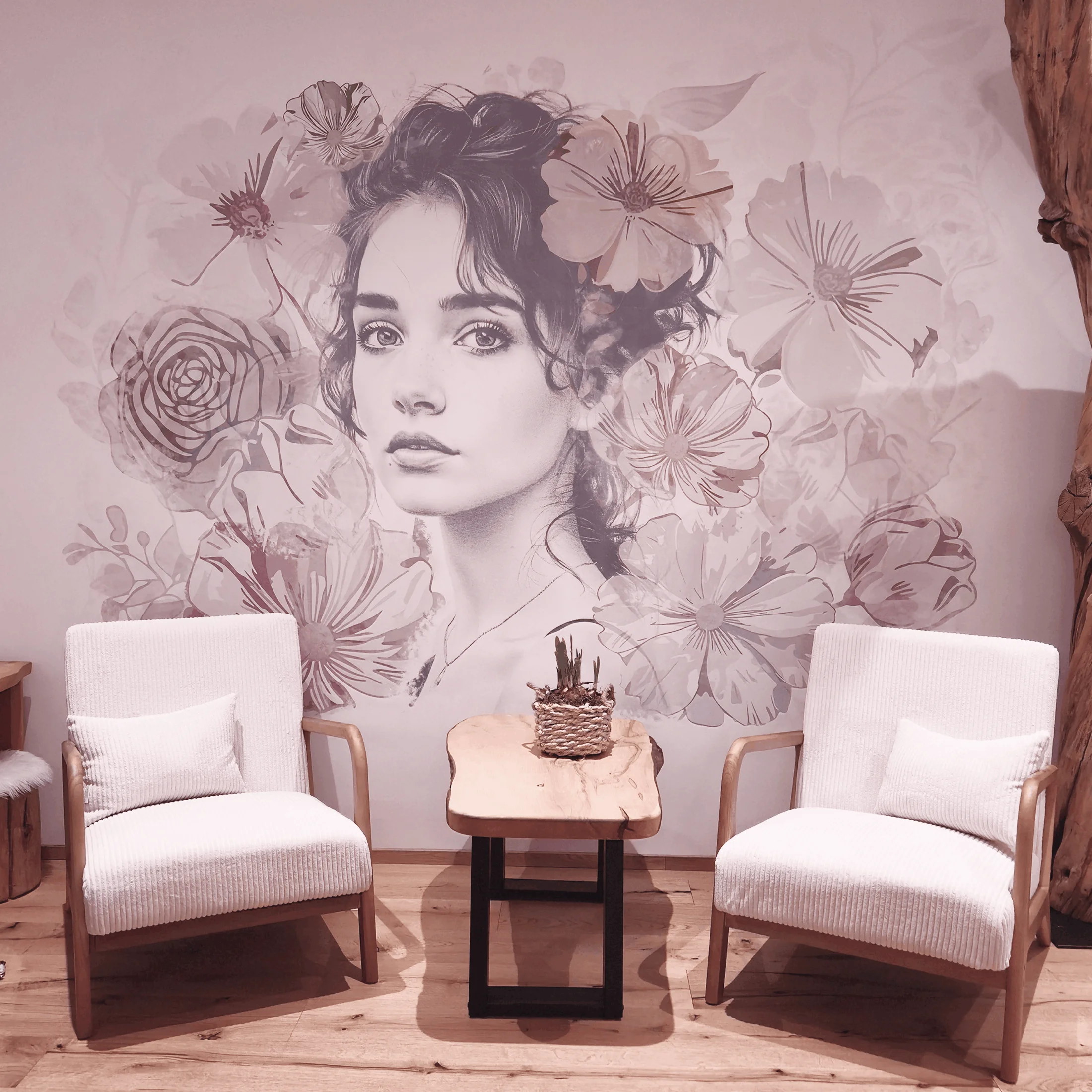 Zwei weiße Sesseln mit Holzarmelehnen und Kissen, mit einem Holztisch dazwischen und einer Wand mit einem großen Porträt einer Frau umgeben von Blumen im Hintergrund.