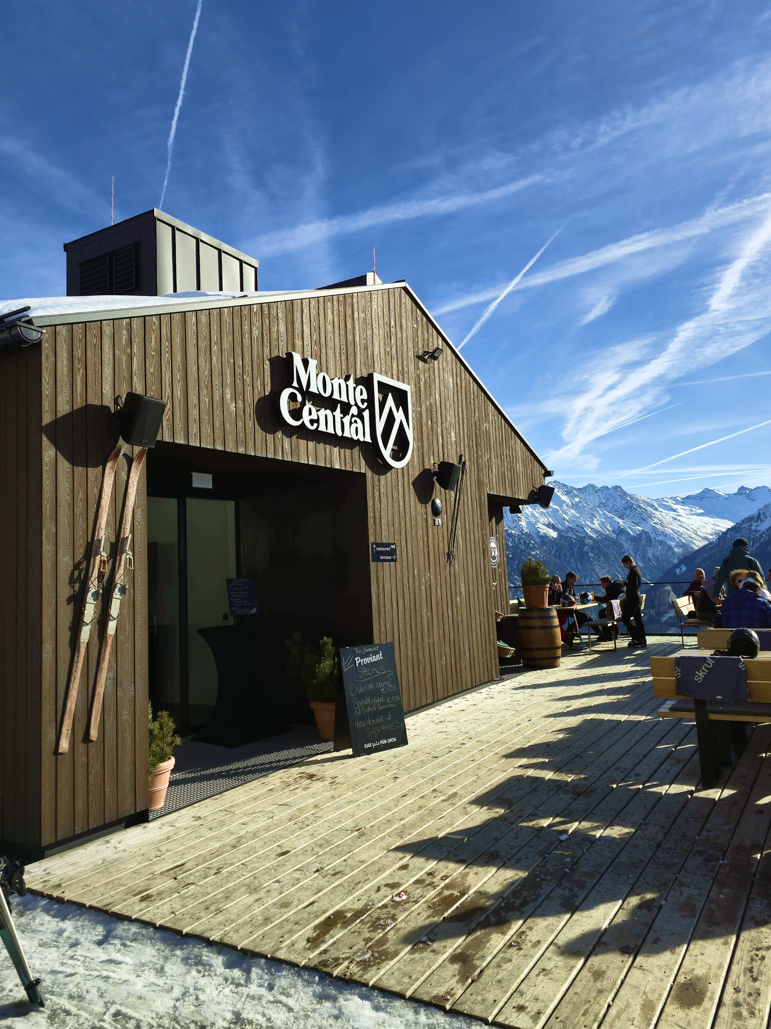 Monte Central Holzhütte mit Terrasse und Gästen vor schneebedeckten Bergen unter blauem Himmel.
