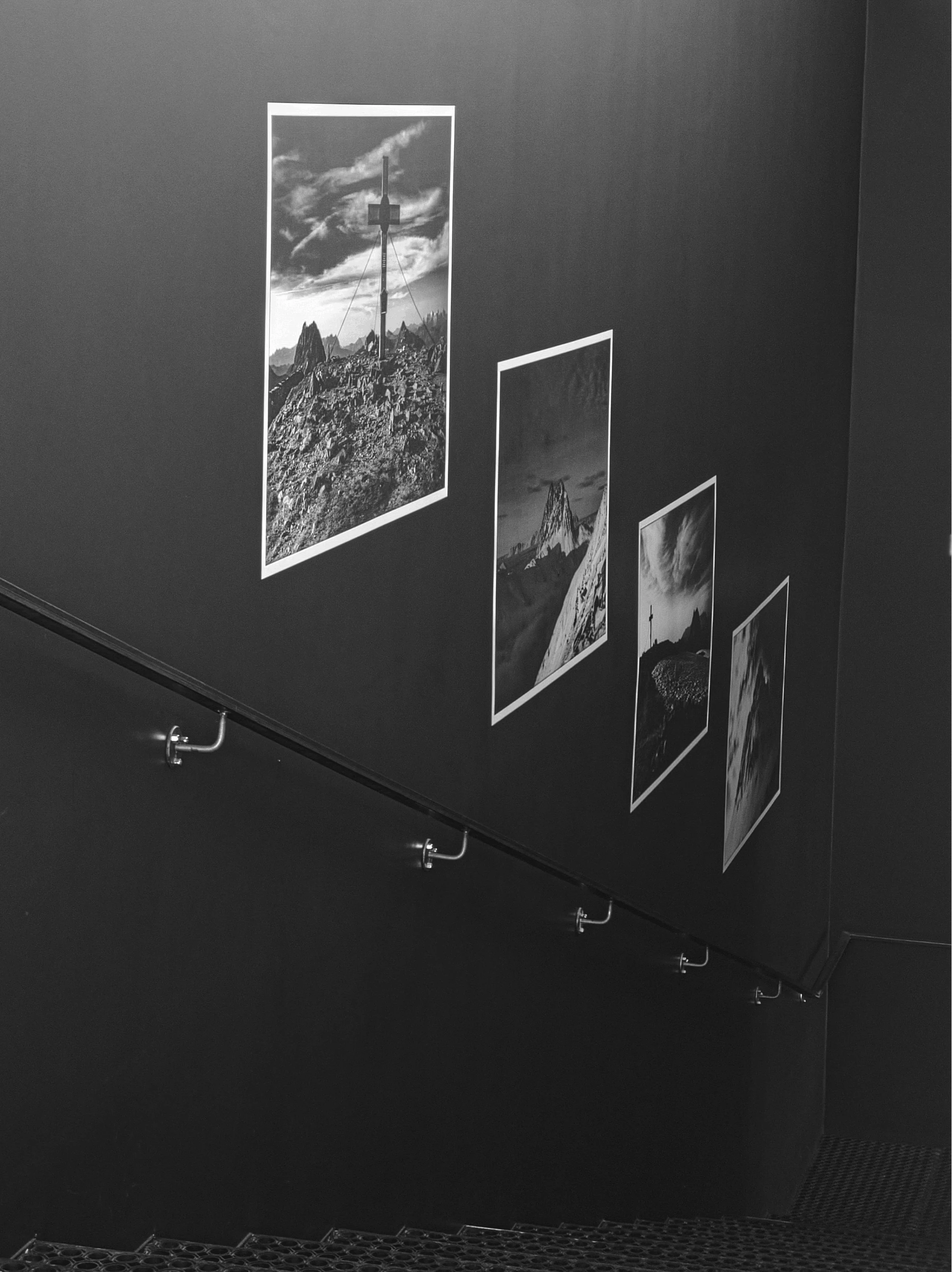 Schwarz-weiß-Foto einer Treppe mit dunklen Wänden, an denen vier gerahmte Bergpanorama-Bilder hängen.