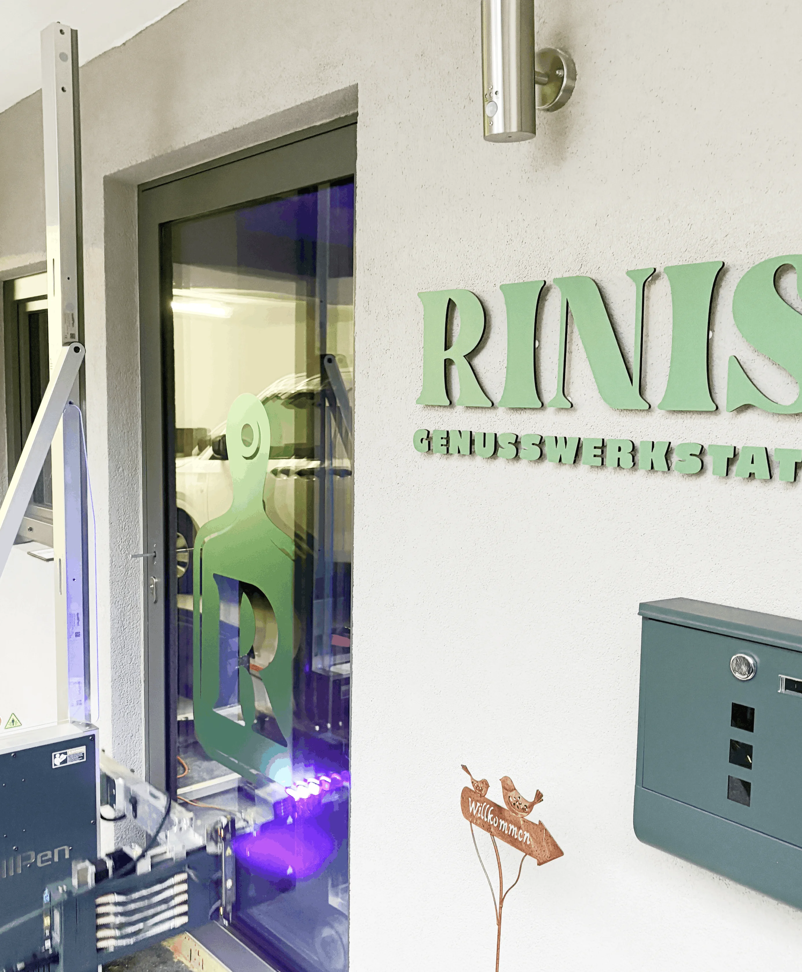 Eingangstür mit grünem Logo und Schriftzug 'RINIS Genusswerkstatt' an weißer Wand, daneben grüner Briefkasten und kleines Willkommensschild mit Vögeln.