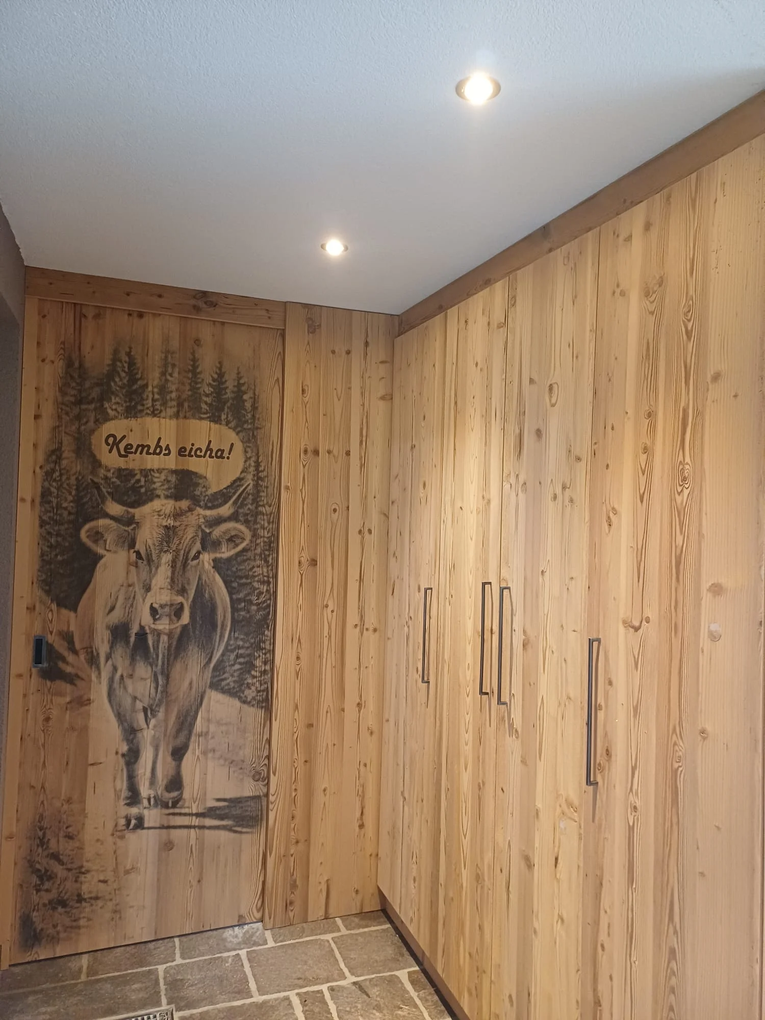 Holztür mit einem aufgedruckten Bild einer Kuh im Wald und der Aufschrift ‚Kembs eicha!‘, daneben eine Wand mit Holzschränken und Metallgriffen.