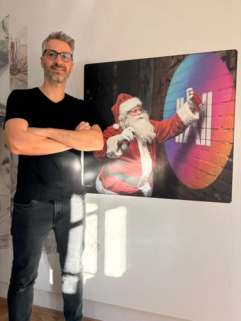 Ein Mann mit Brille, schwarzem T-Shirt und verschränkten Armen steht neben einem Wandbild von Santa Claus, der mit Sprühdosen ein buntes Wandsignet gestaltet.