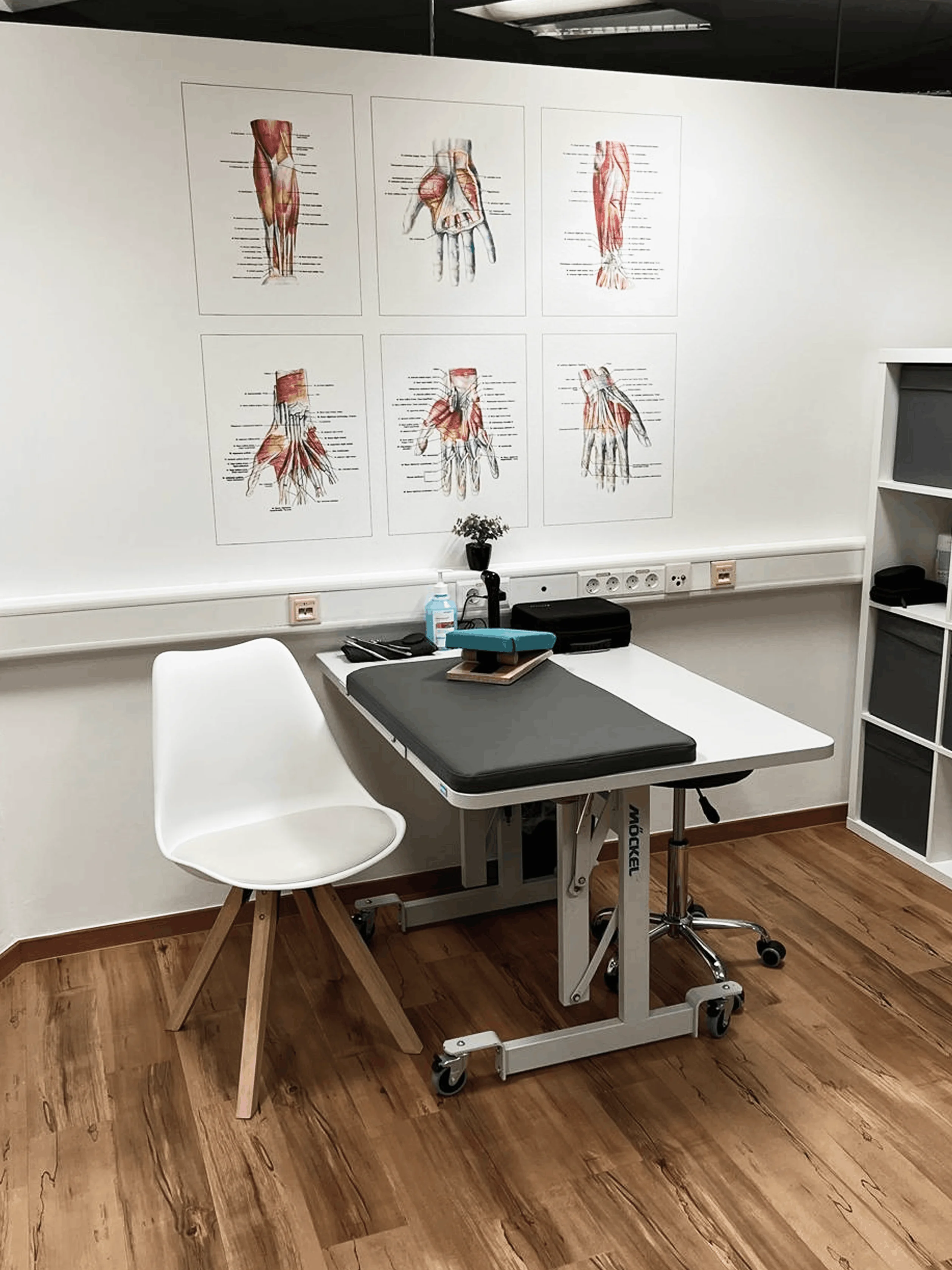 Behandlungsraum mit weißem Stuhl, verstellbarem Untersuchungstisch, anatomischen Handmuskel-Diagrammen an der Wand und medizinischen Utensilien.
