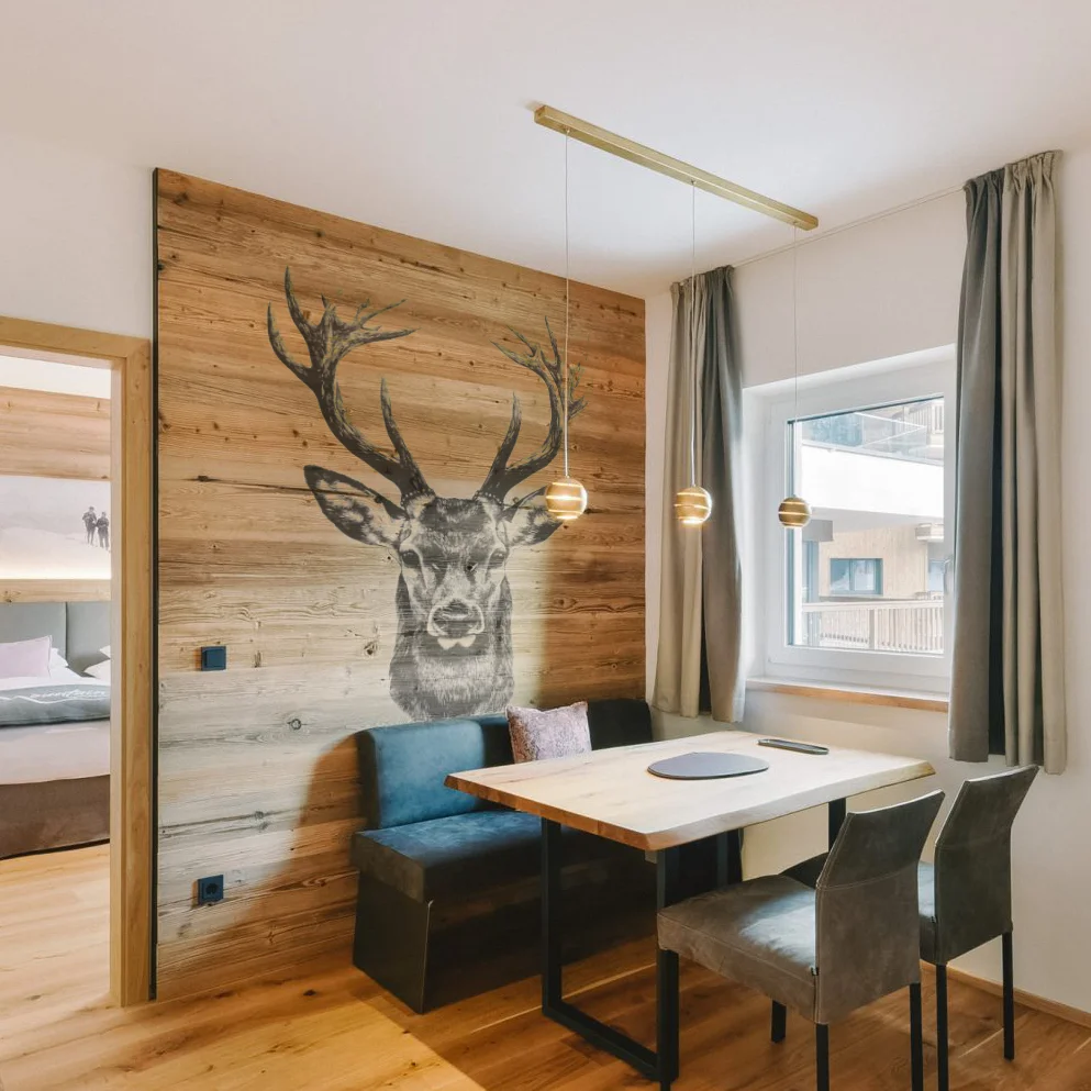 Essbereich mit Holztisch, Sesseln und blauer Bank vor einer Holzwand mit großem Hirschkopf-Motiv, neben Fenster mit langen Vorhängen.