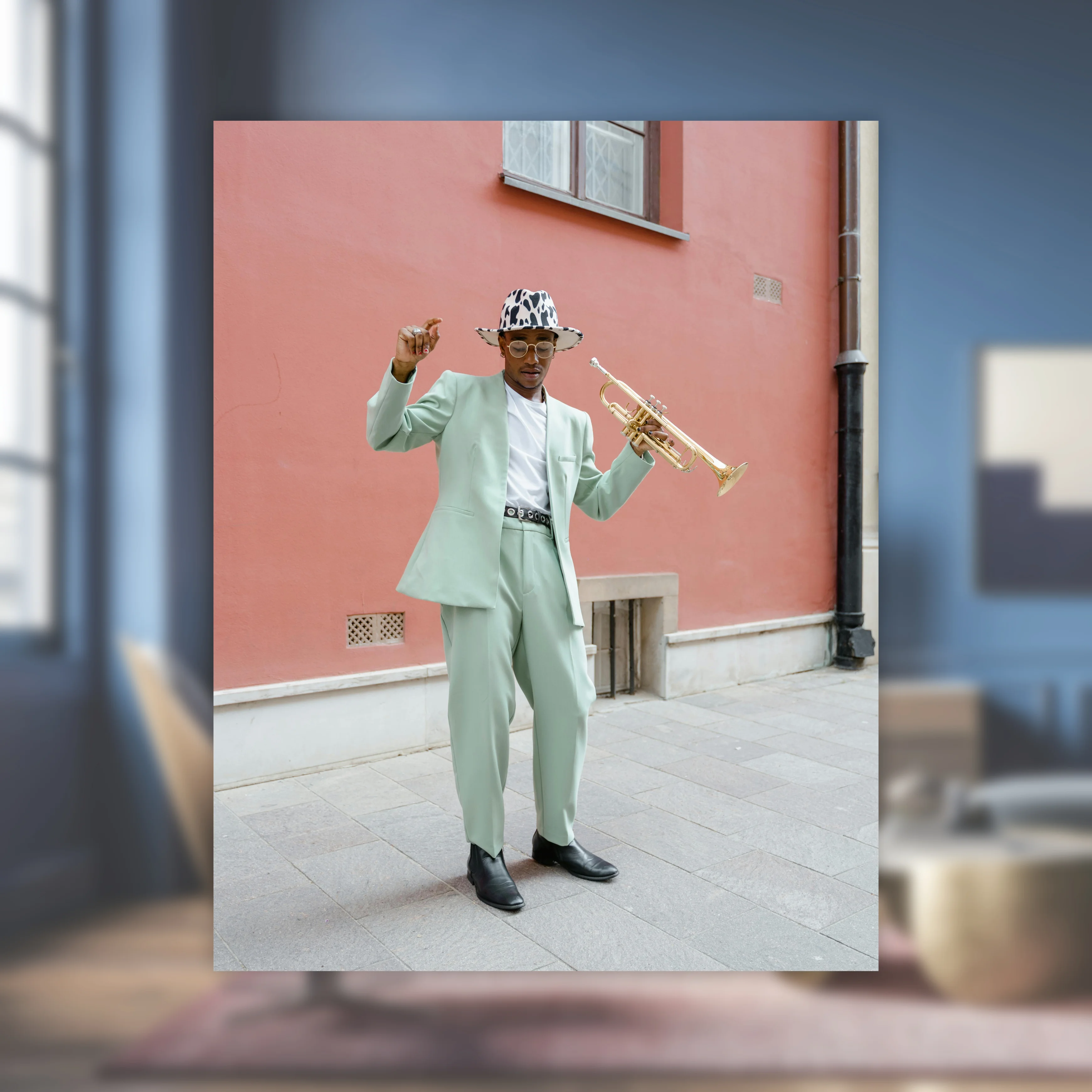 Mann in einem hellgrünen Anzug und Hut mit Kuhmuster, der eine Trompete hält und vor einer rosa Wand posiert.