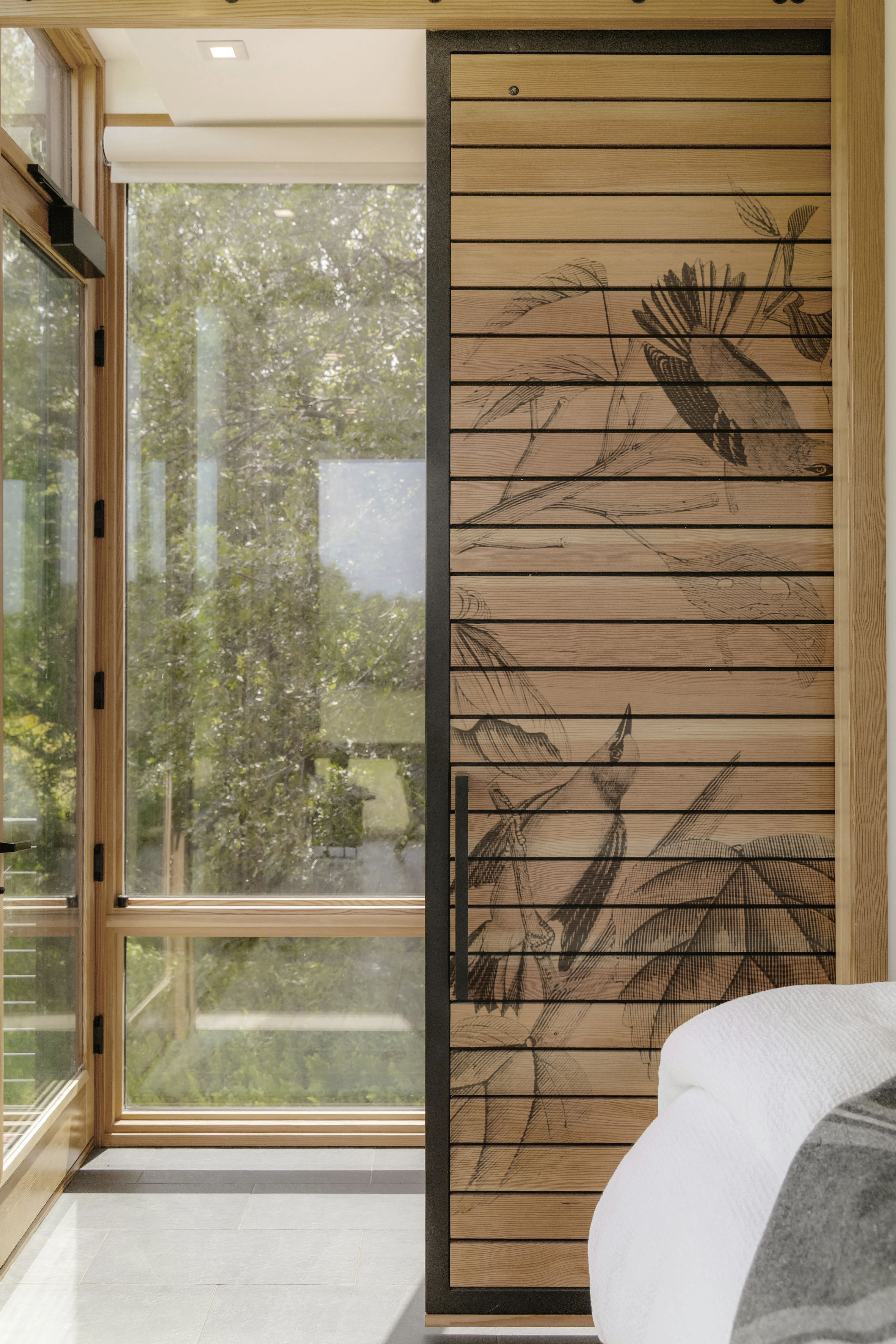 Schiebetür aus Holz mit schwarzer Vogel- und Pflanzenzeichnung neben einem großen Fenster mit Blick ins Grüne.