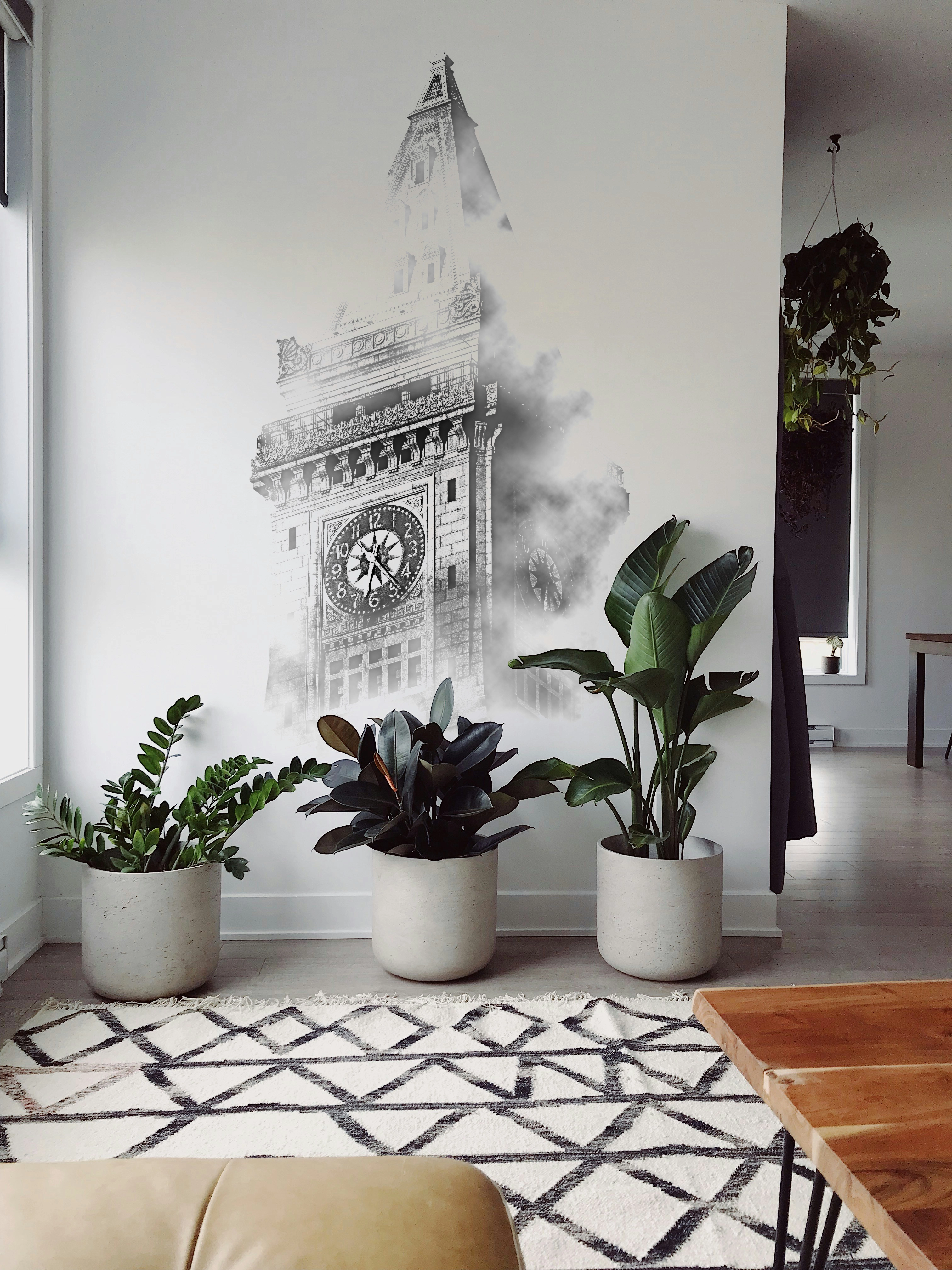 Drei große Pflanzen in Töpfen stehen vor einer Wand mit einer künstlerischen Uhrturmzeichnung in einem modernen Wohnzimmer.