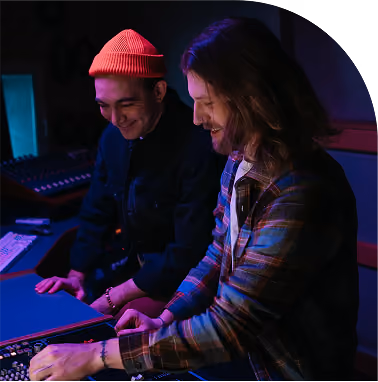 Two men smiling and working on a mixing console in a dimly lit studio with blue and purple lighting.