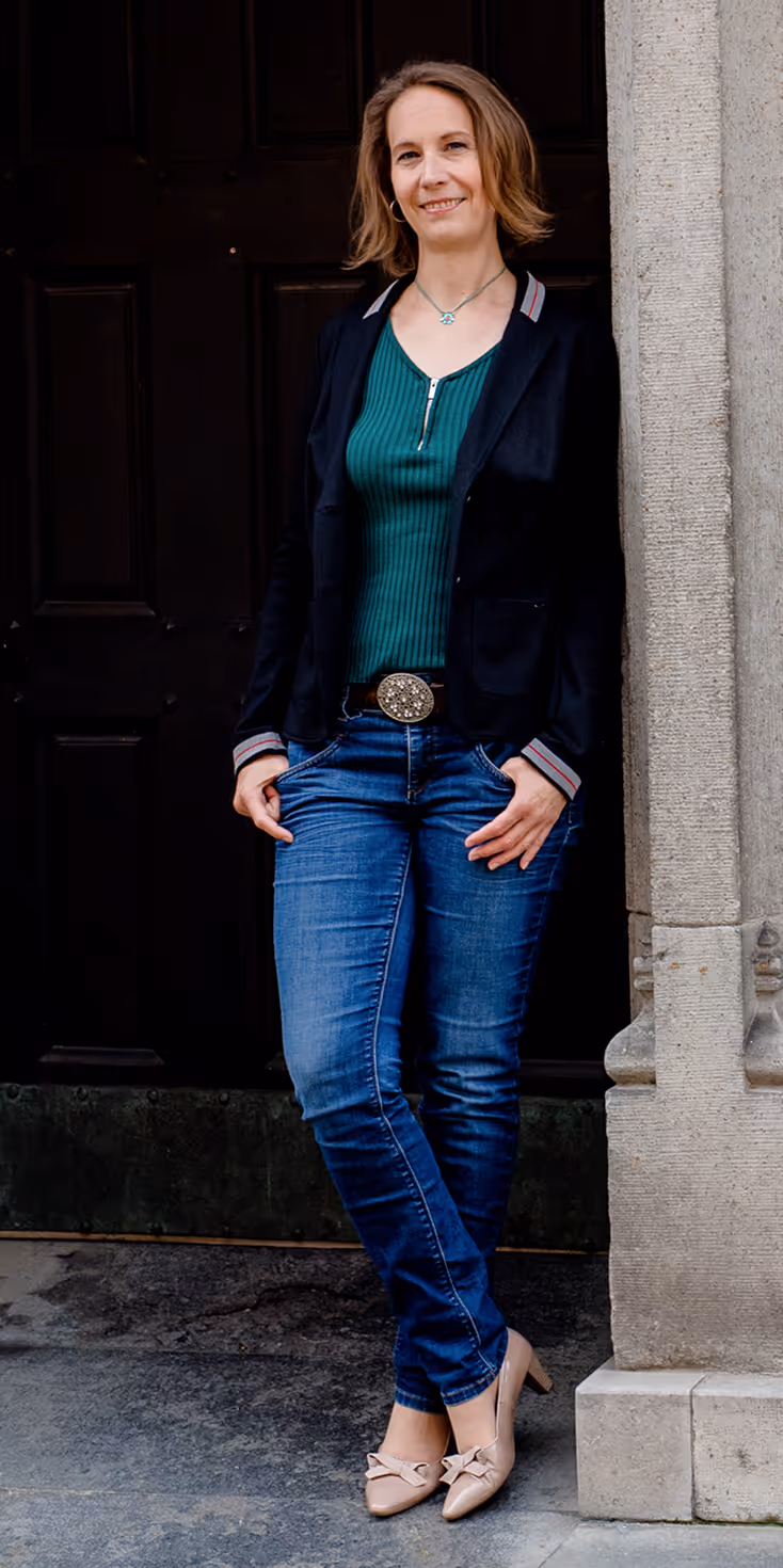 Frau lehnt an einem steinernen Türrahmen, trägt blau-grünes Oberteil, schwarzen Blazer, Jeans und beige Schuhe mit Absatz.