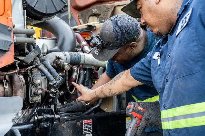 Two technicians perform a coolant leak inspection near turbo piping and hoses on a heavy-duty diesel engine.