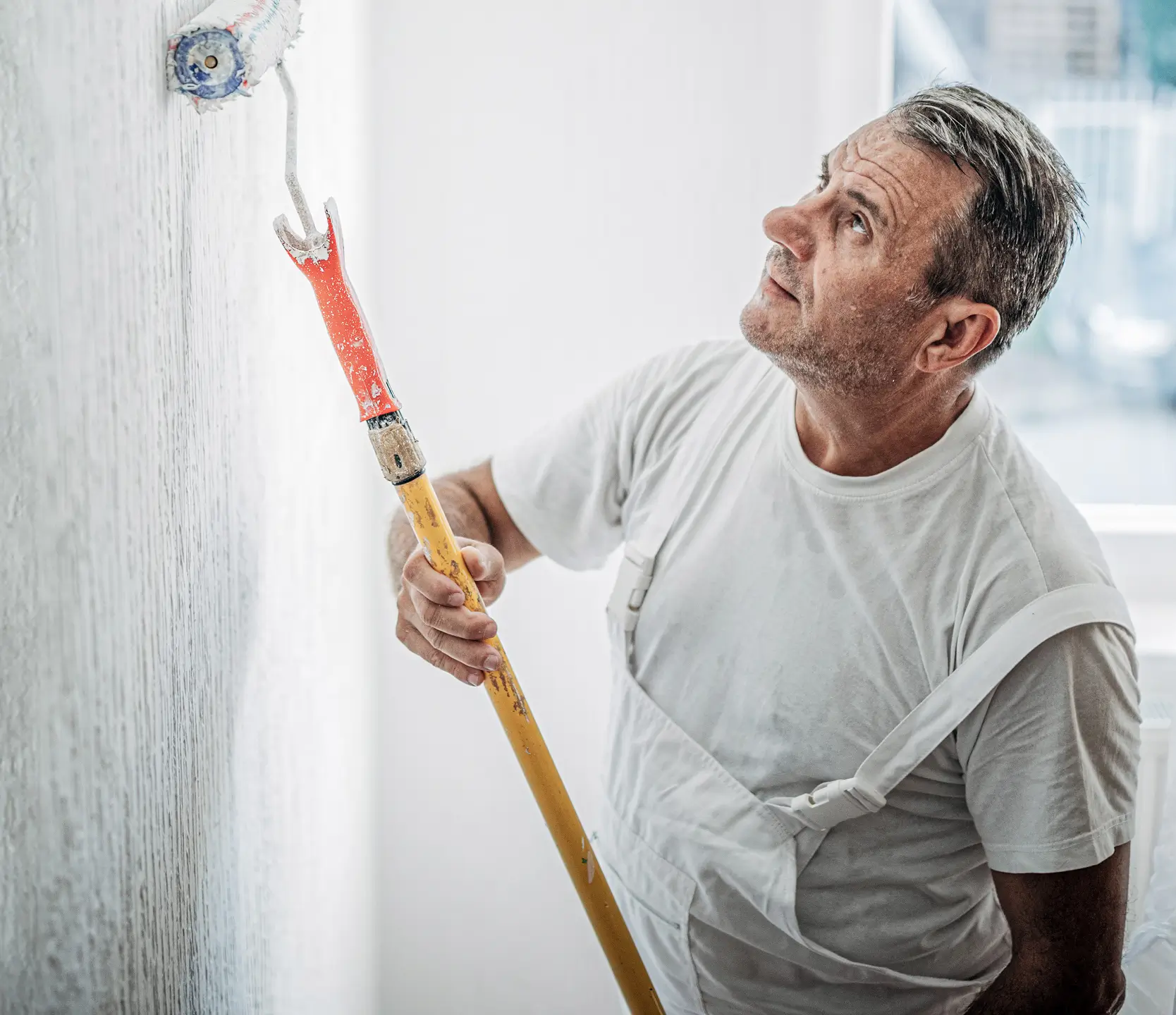 A decorator paints a white wall using a long-handled roller, focused on achieving a smooth, even finish.