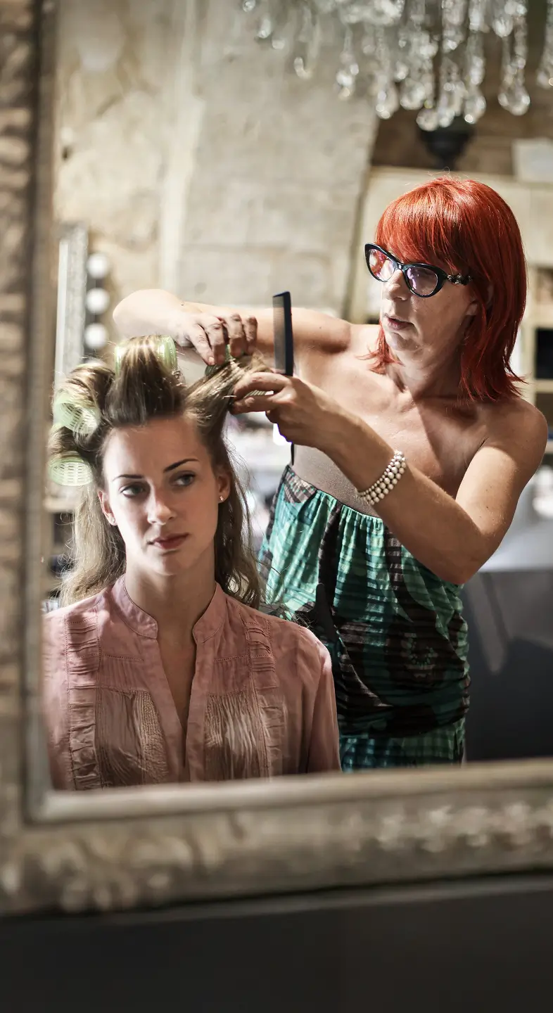 A hairstylist styles a client’s hair in front of a large salon mirror, focused and professional in a well-lit space.