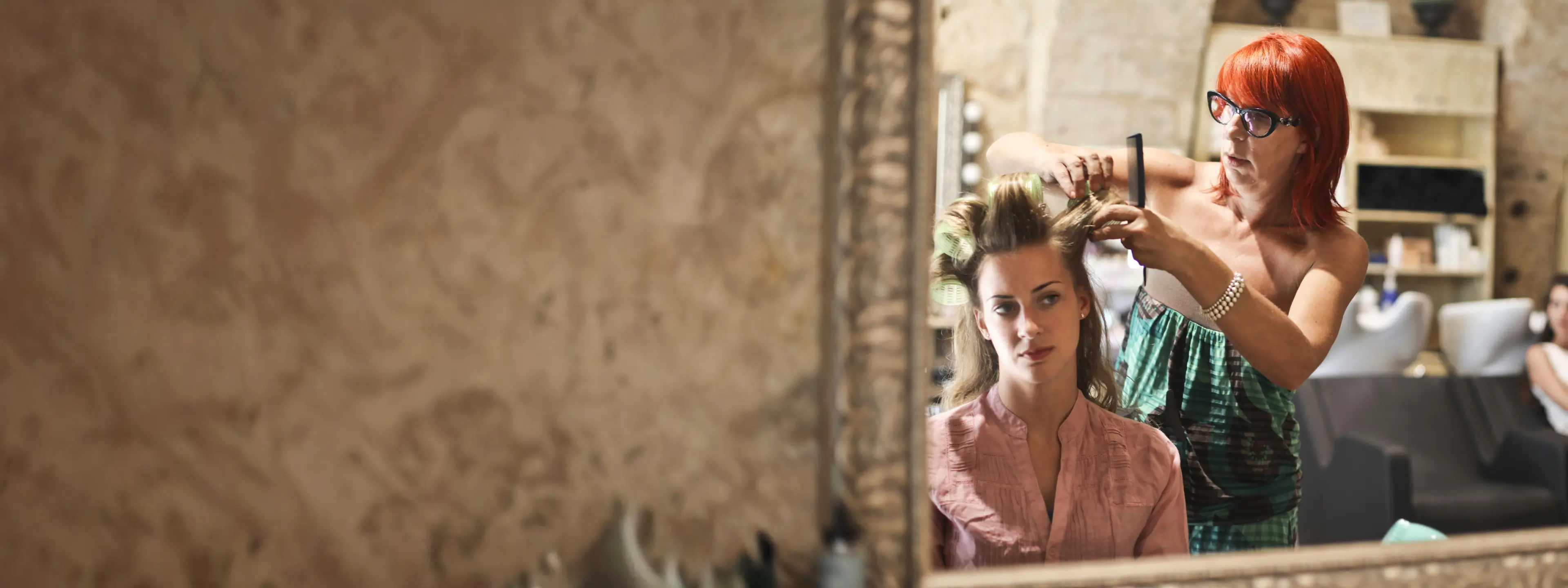A hairstylist styles a client’s hair in front of a large salon mirror, focused and professional in a well-lit space.