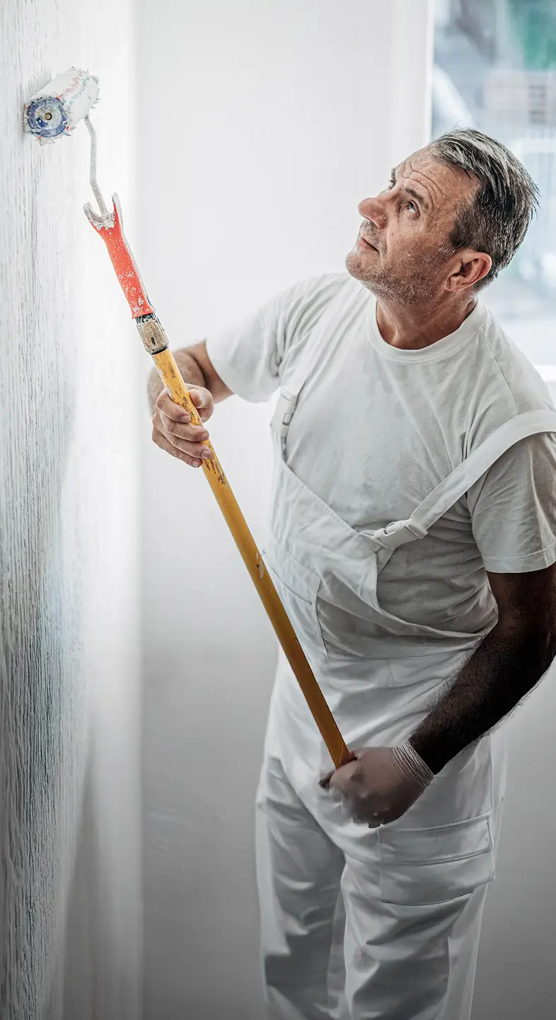 A decorator paints a white wall using a long-handled roller, focused on achieving a smooth, even finish.