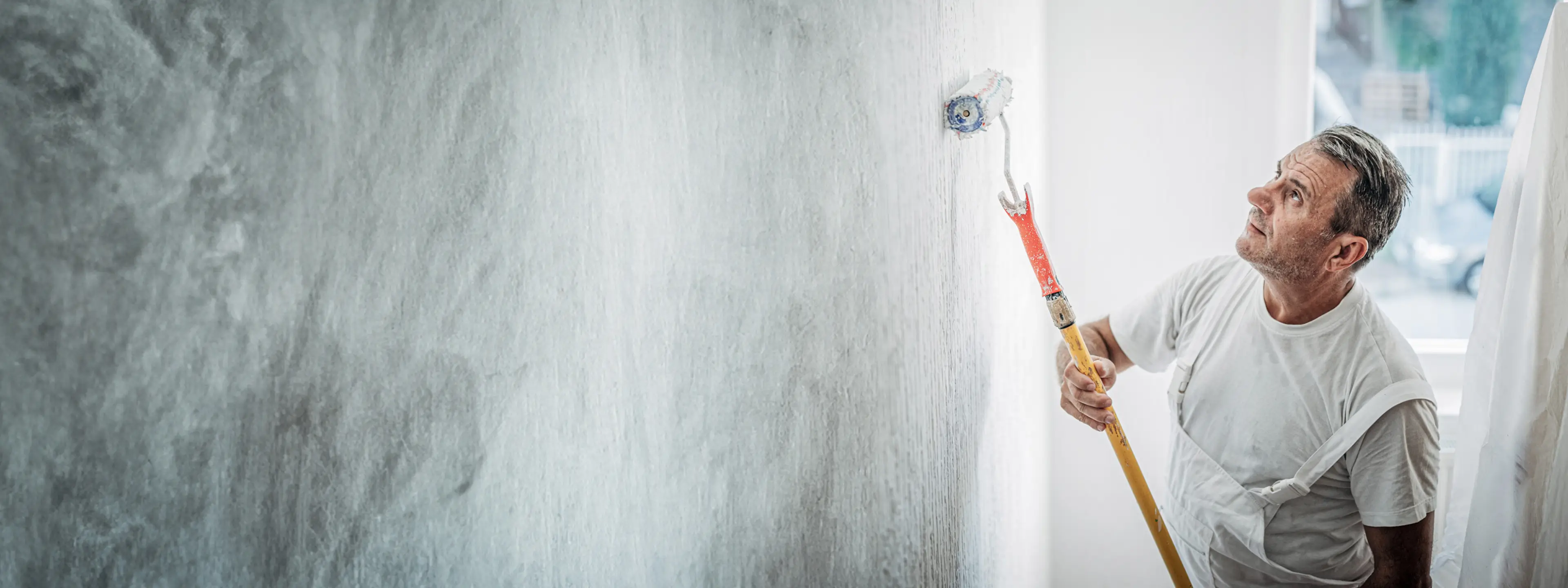 A decorator paints a white wall using a long-handled roller, focused on achieving a smooth, even finish.