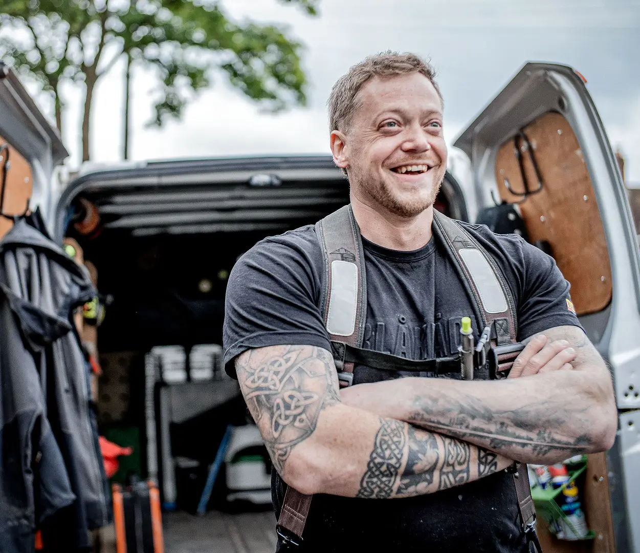 A confident tradesperson stands in front of an open van fully stocked with tools, smiling with arms crossed, ready for the day’s work.